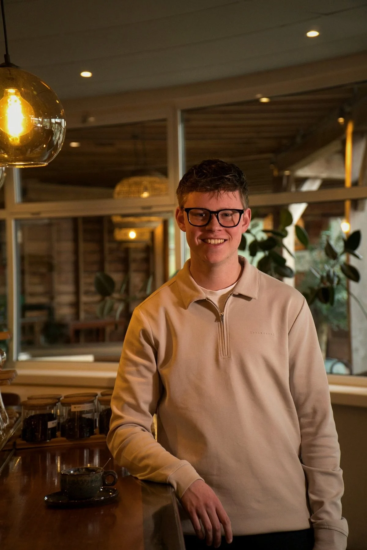 Een jonge man met bril en een beige trui, lachend in een gezellige binnenruimte met planten en houten interieur.