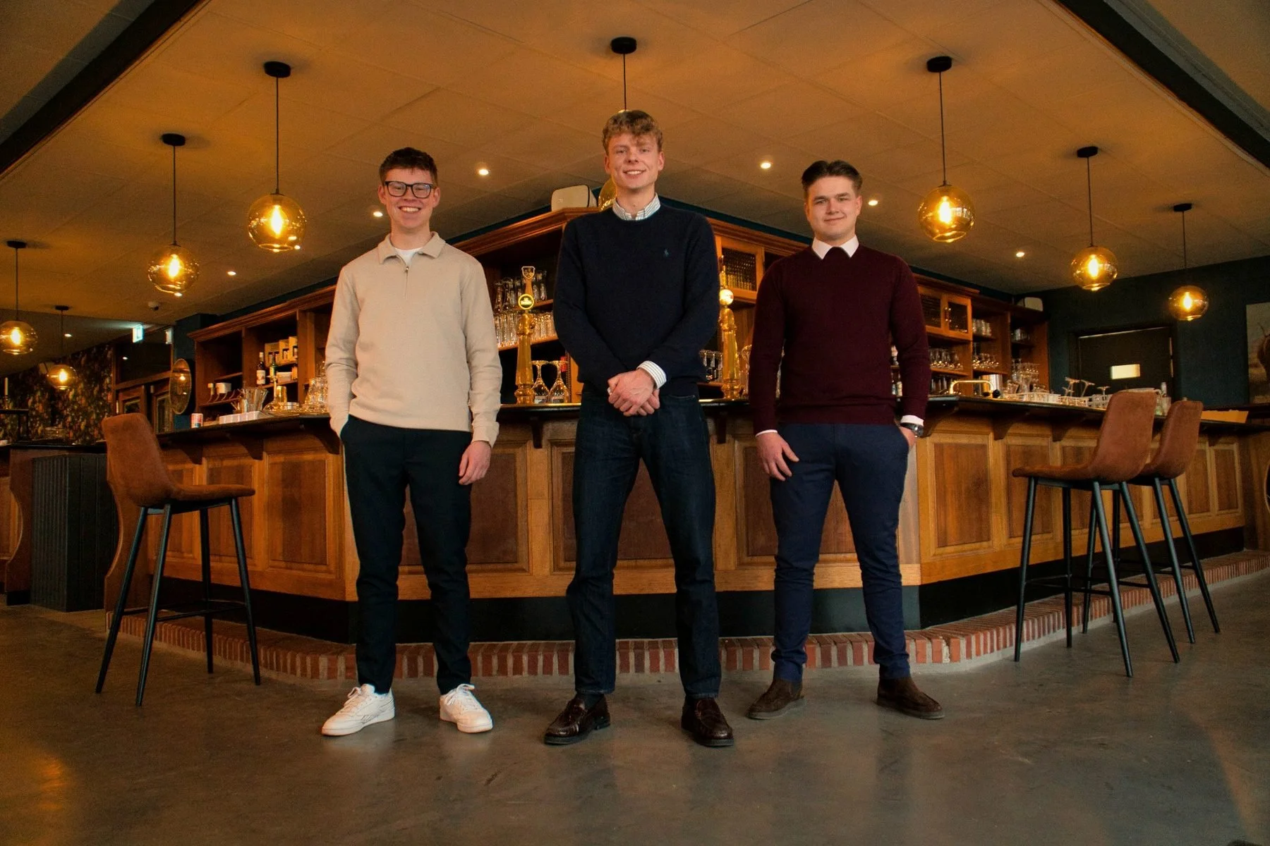 Drie jonge mannen staan in een bar, lachend en poseerend voor de camera.