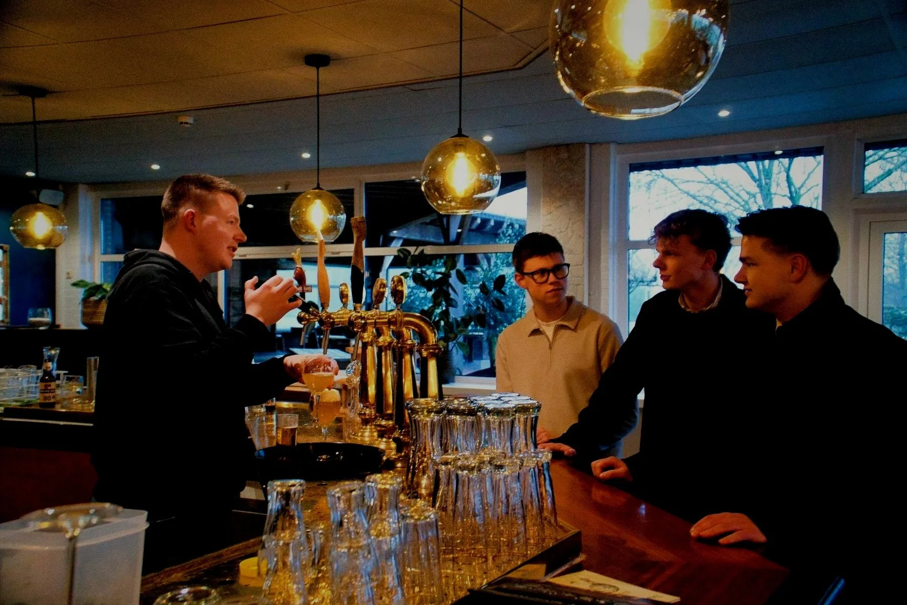 Een bartender schenkt een drankje in een glas terwijl drie mannen aan de bar staan en met hem praten in een restaurant of café.