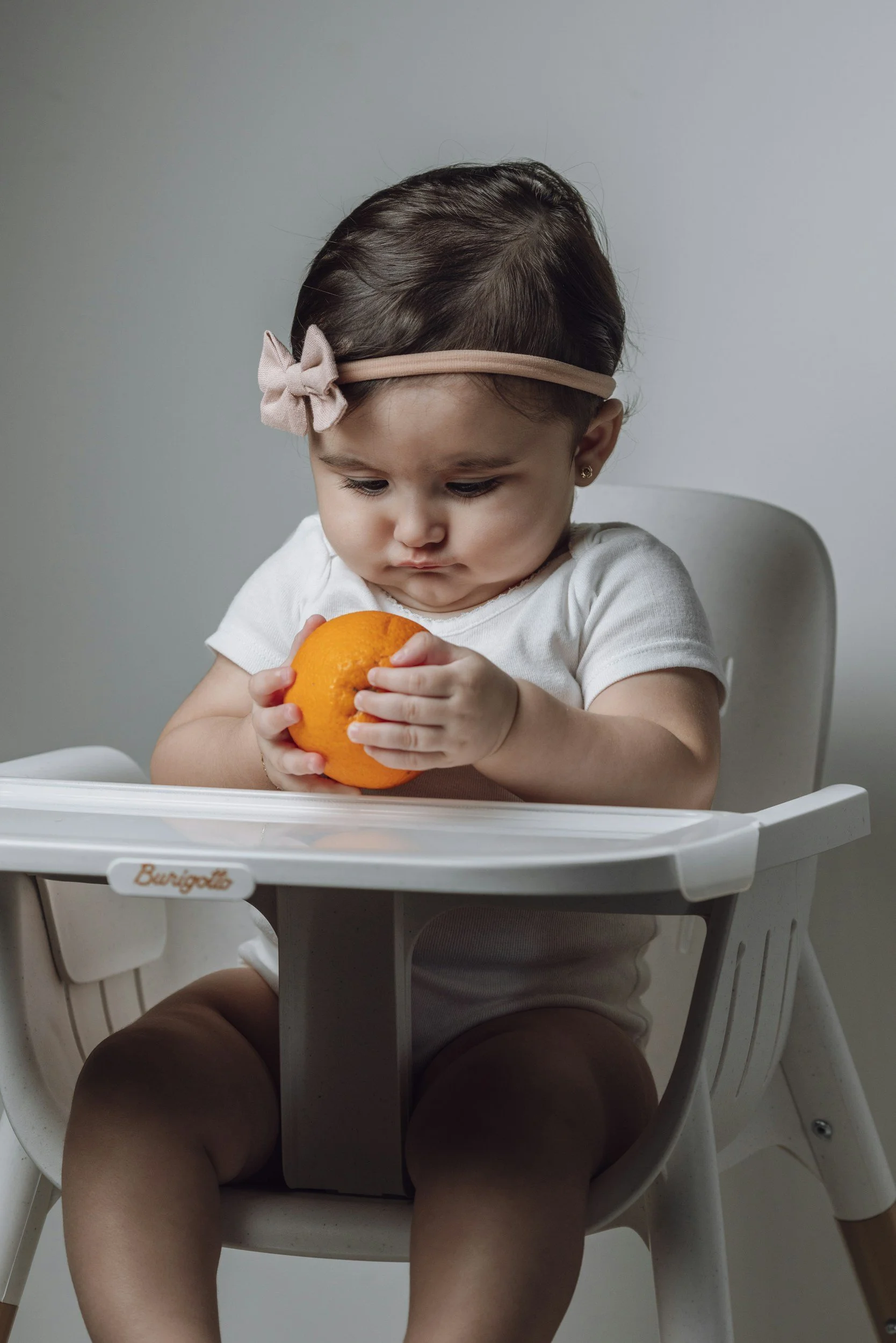 A young girl with dark hair, wearing a pink headband with a bow, sitting in a white high chair, holding and looking at an orange fruit with a focused expression.