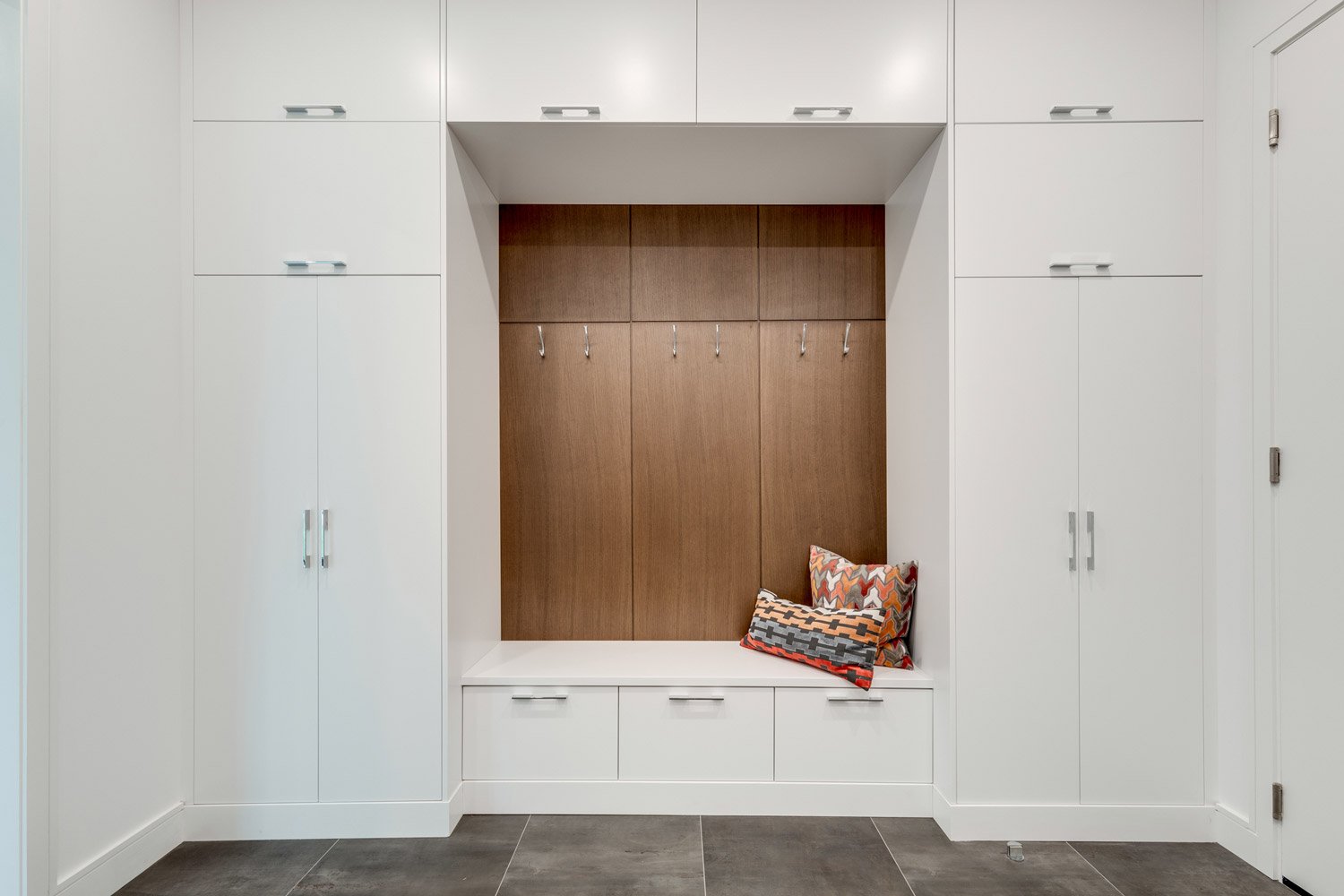 Modern built-in storage unit with white cabinets, a wood-paneled center area with hooks, and a bench with two decorative pillows.