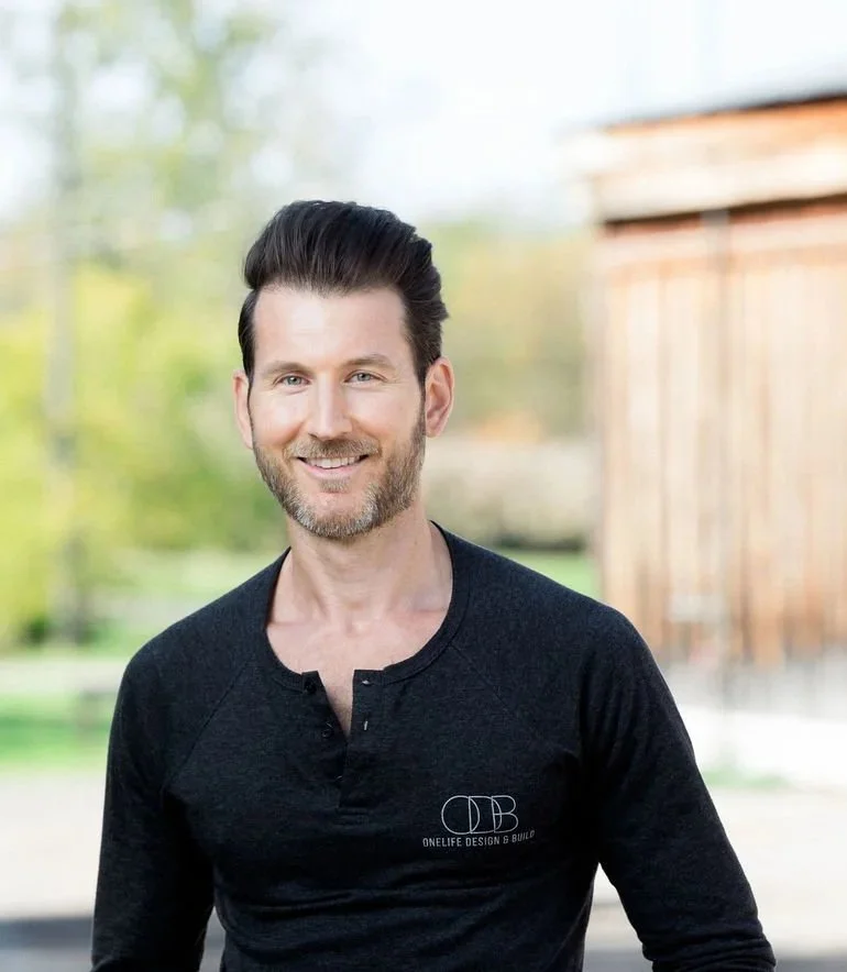 A smiling man with dark, styled hair and a beard, wearing a black long-sleeve shirt with a logo that reads "ONE LIFE DESIGN & BUILD," standing outdoors with a blurred background of trees and a wooden building.