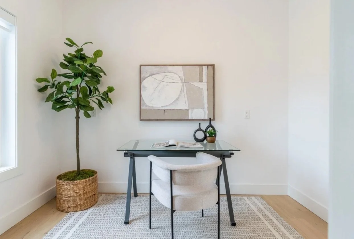 Minimalist home office with a glass desk, white upholstered chair, large potted plant, abstract art on the wall, and decorative black vases, on a light-colored rug.