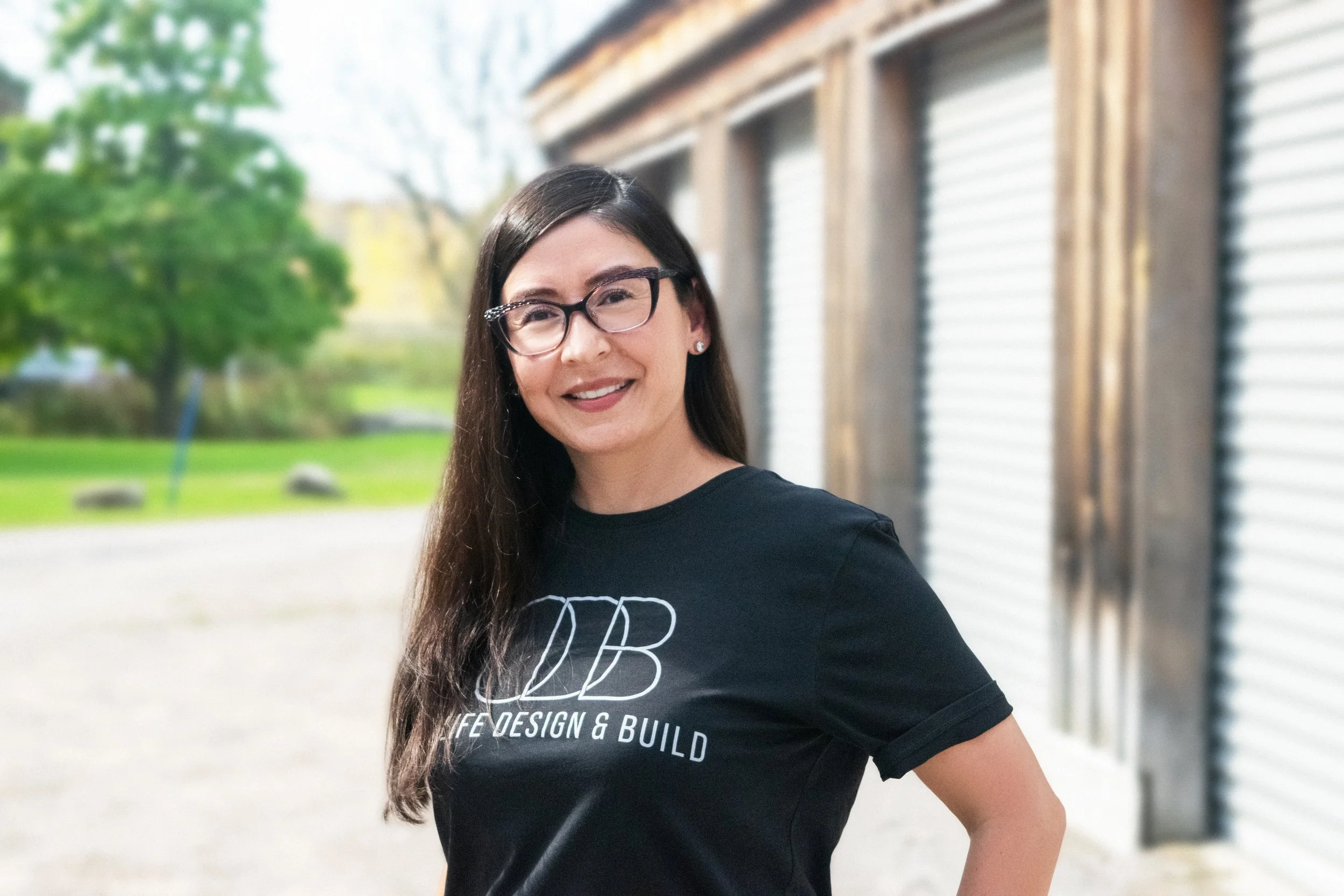 A woman with long dark hair, glasses, and earrings standing outdoors, smiling at the camera, wearing a black t-shirt with 'LIFE DESIGN & BUILD' written on it.