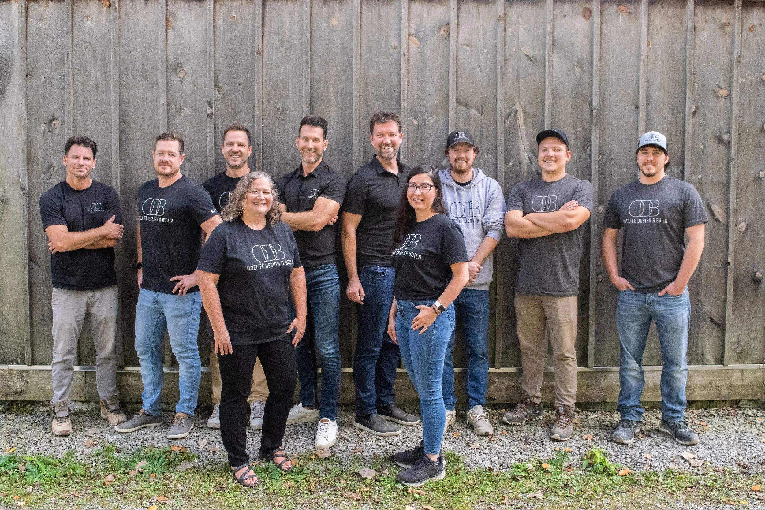 A group of eleven people standing outdoors in front of a wooden fence, some wearing matching black or grey T-shirts with the logo 'OB' and the words 'OneLife Design & Build,' smiling at the camera.