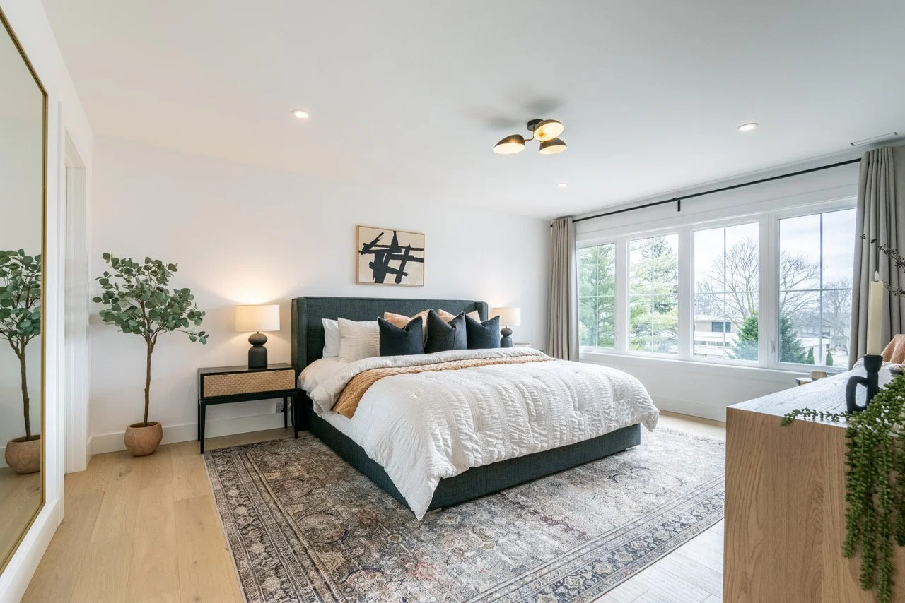 Modern bedroom with large window view, king-sized bed with black headboard and white bedding, two bedside lamps, artwork above the bed, plants, and wooden furniture.