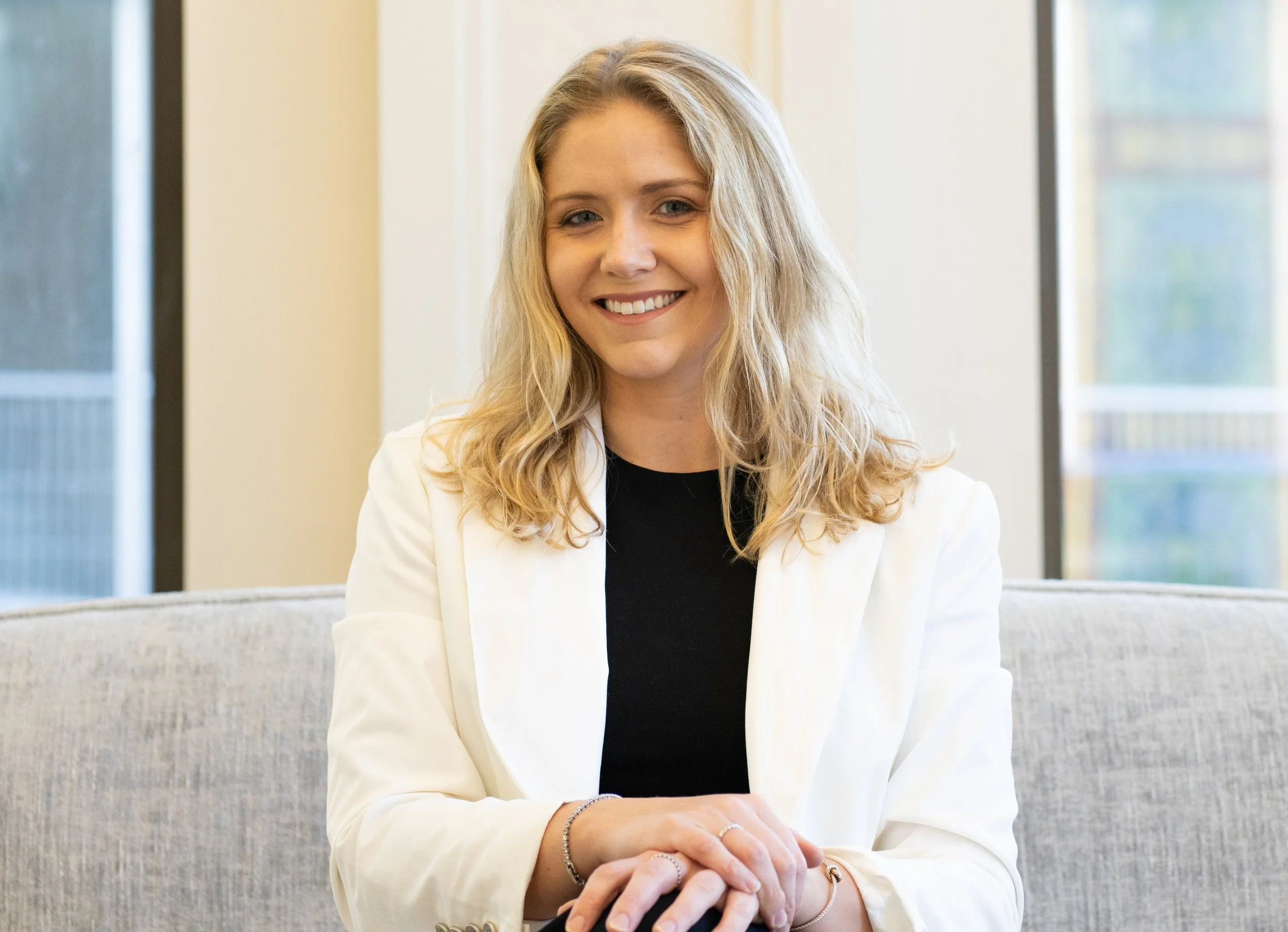 A smiling woman with blonde, wavy hair sitting on a light-colored sofa in front of a window with a view of trees.