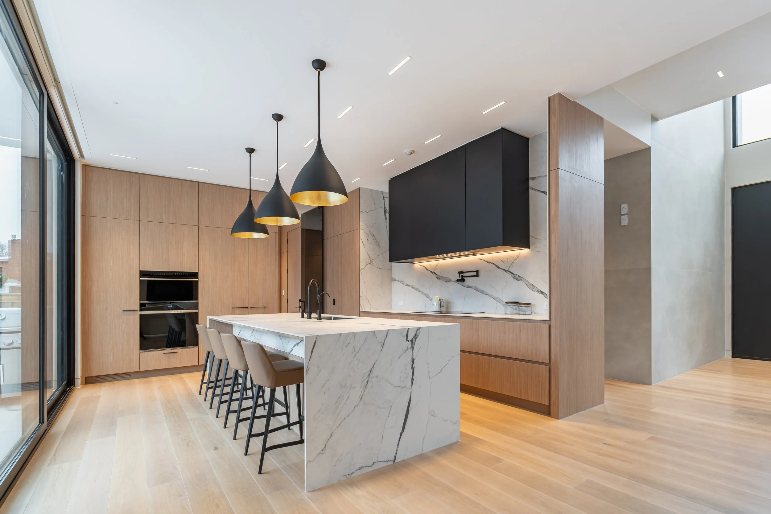 Modern kitchen with marble island, wooden cabinets, black upper cabinets, and a wooden dining table with beige chairs, illuminated by three black pendant lights.