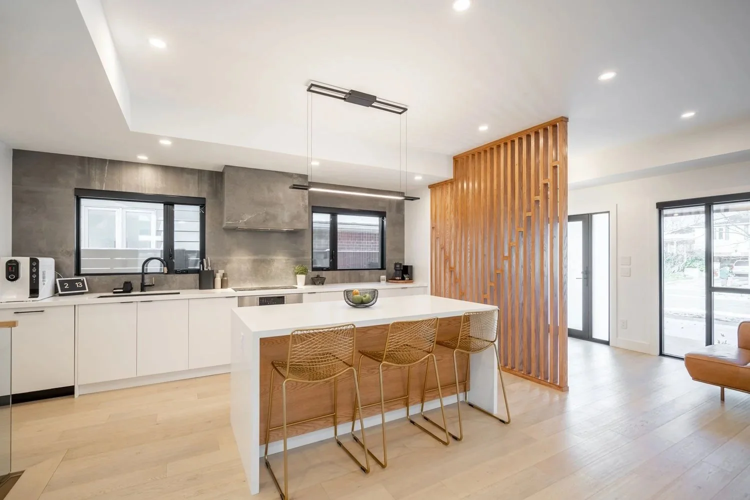 Modern kitchen and living area with a white island, bar stools, gray textured backsplash, black window frames, a wooden slat room divider, and large glass sliding doors.