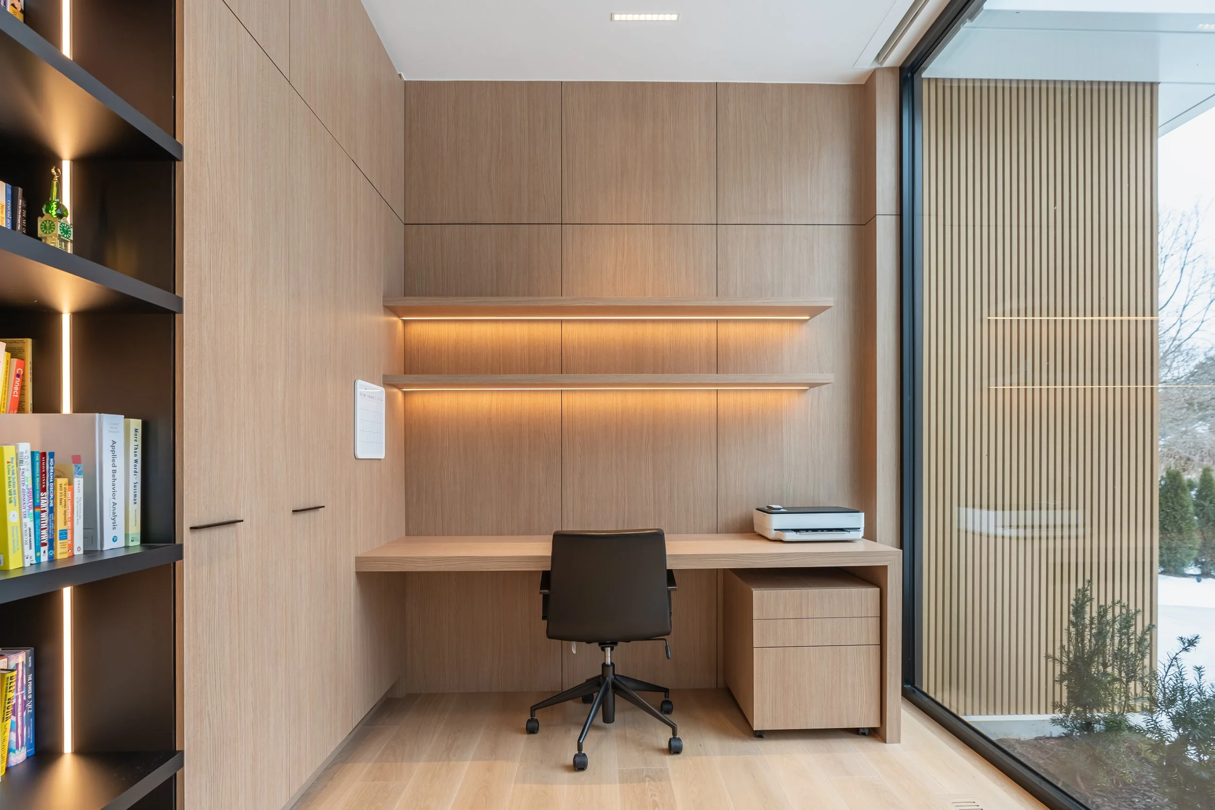Modern home office with built-in wooden desk, black swivel chair, and wooden shelves with LED lighting, next to large window with wood slats outside.