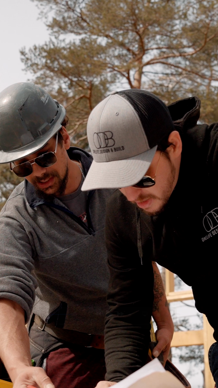 Two construction workers, one wearing a gray hard hat and sunglasses, and the other wearing a black and gray baseball cap, are focused on a construction plan or blueprint outdoors with trees in the background.