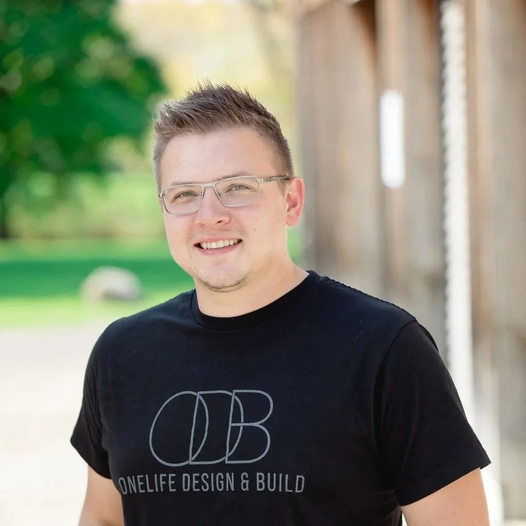 A young man with glasses, short light brown hair, and a slight beard, smiling outdoors next to a wooden structure, wearing a black T-shirt with the text 'ONE LIFE DESIGN & BUILD' and a logo.