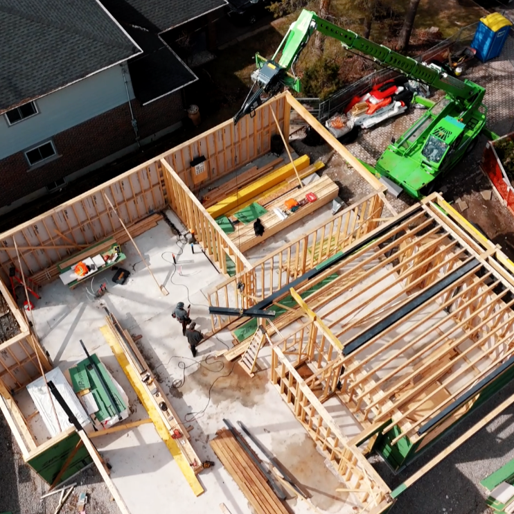 Aerial view of a house under construction with wooden framing, construction workers, and a green crane lifting materials.