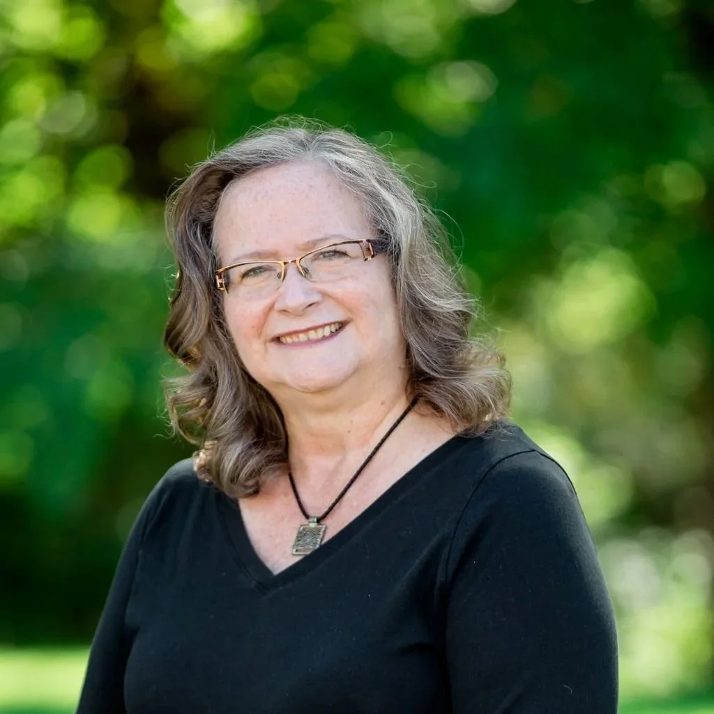A smiling woman with glasses and shoulder-length wavy gray hair, wearing a black top and a necklace, outdoors with a blurred green background.
