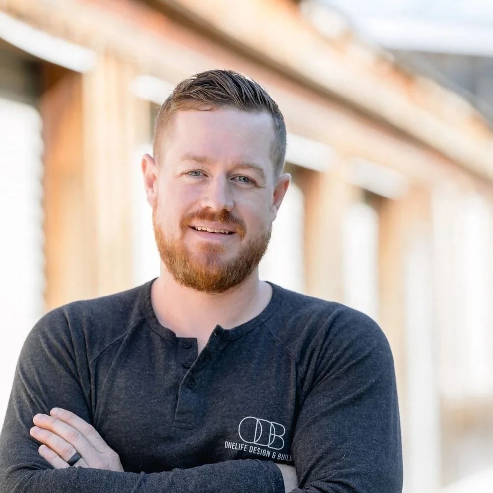 A smiling man with light skin, ginger beard and hair, wearing a black long sleeve shirt with a logo, standing outdoors in front of a wooden building structure.