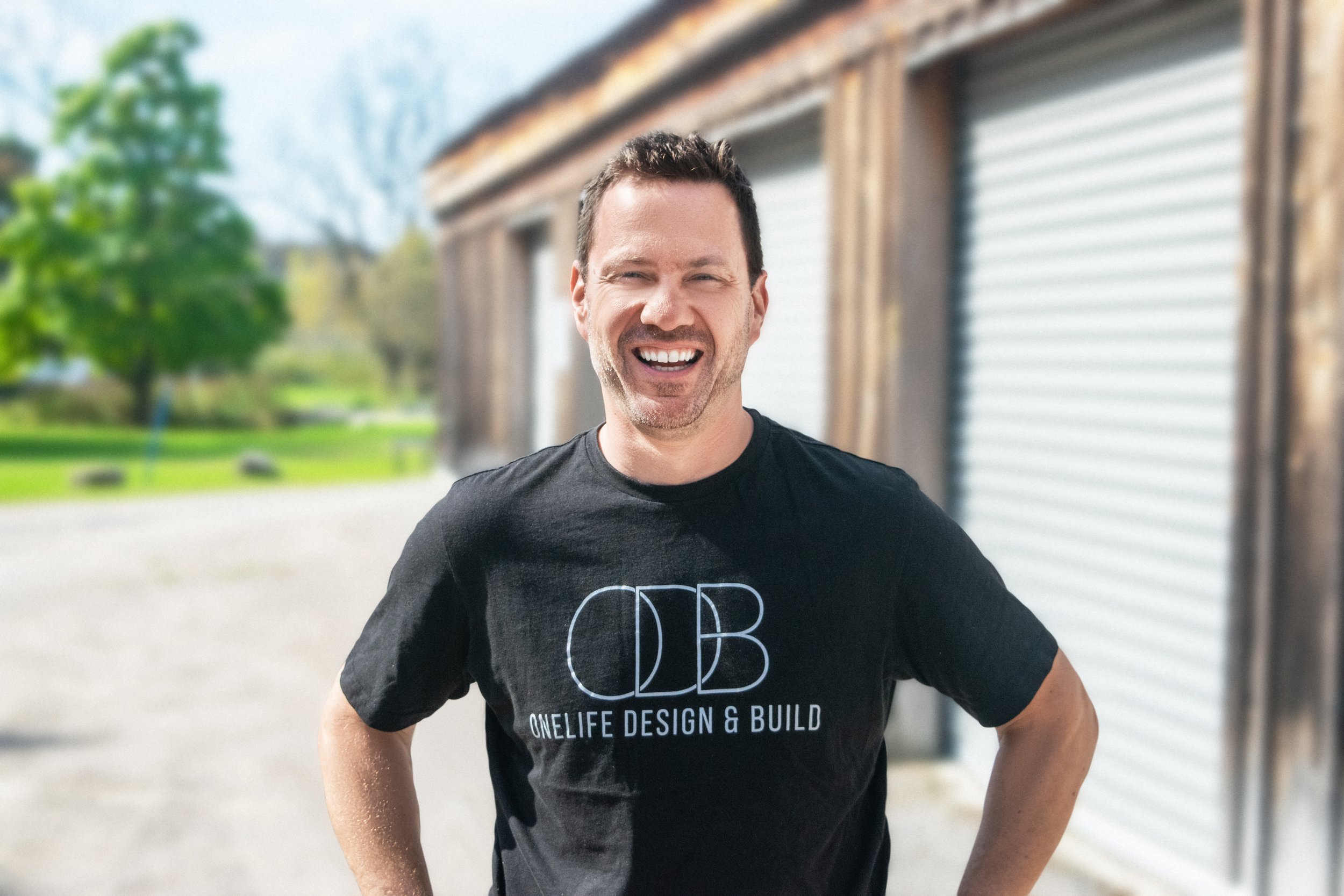 Smiling man standing outdoors in front of a building with trees in the background, wearing a black t-shirt with 'OneLife Design & Build' logo.