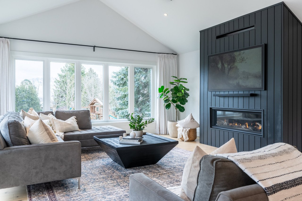 Living room with large windows, gray sofas, a black coffee table with plants, a modern black fireplace with a TV above, and indoor plants.