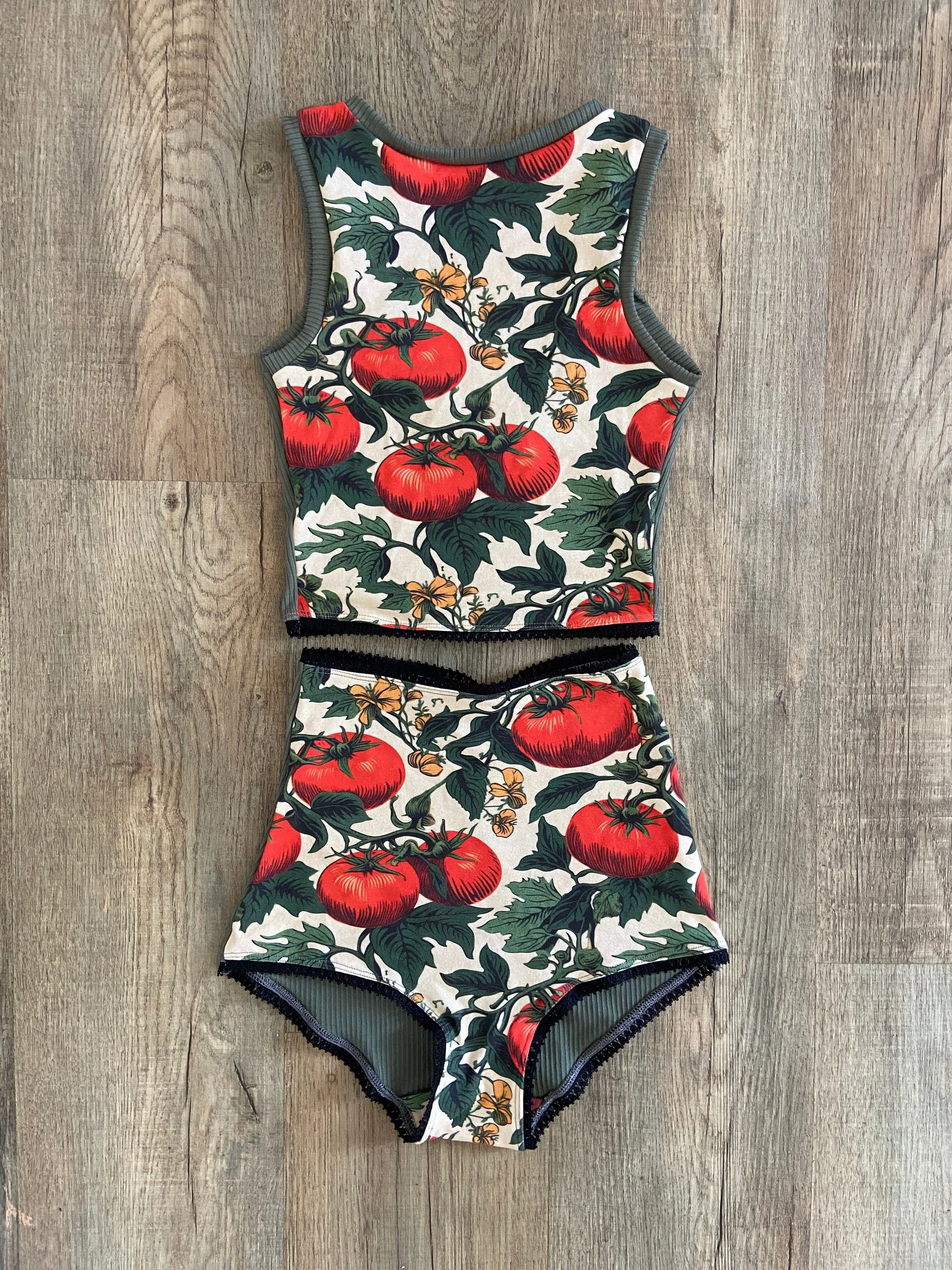 Two-piece swimsuit with a tomato and vine pattern, featuring red tomatoes, green leaves, and orange flowers, laid flat on a wooden surface.