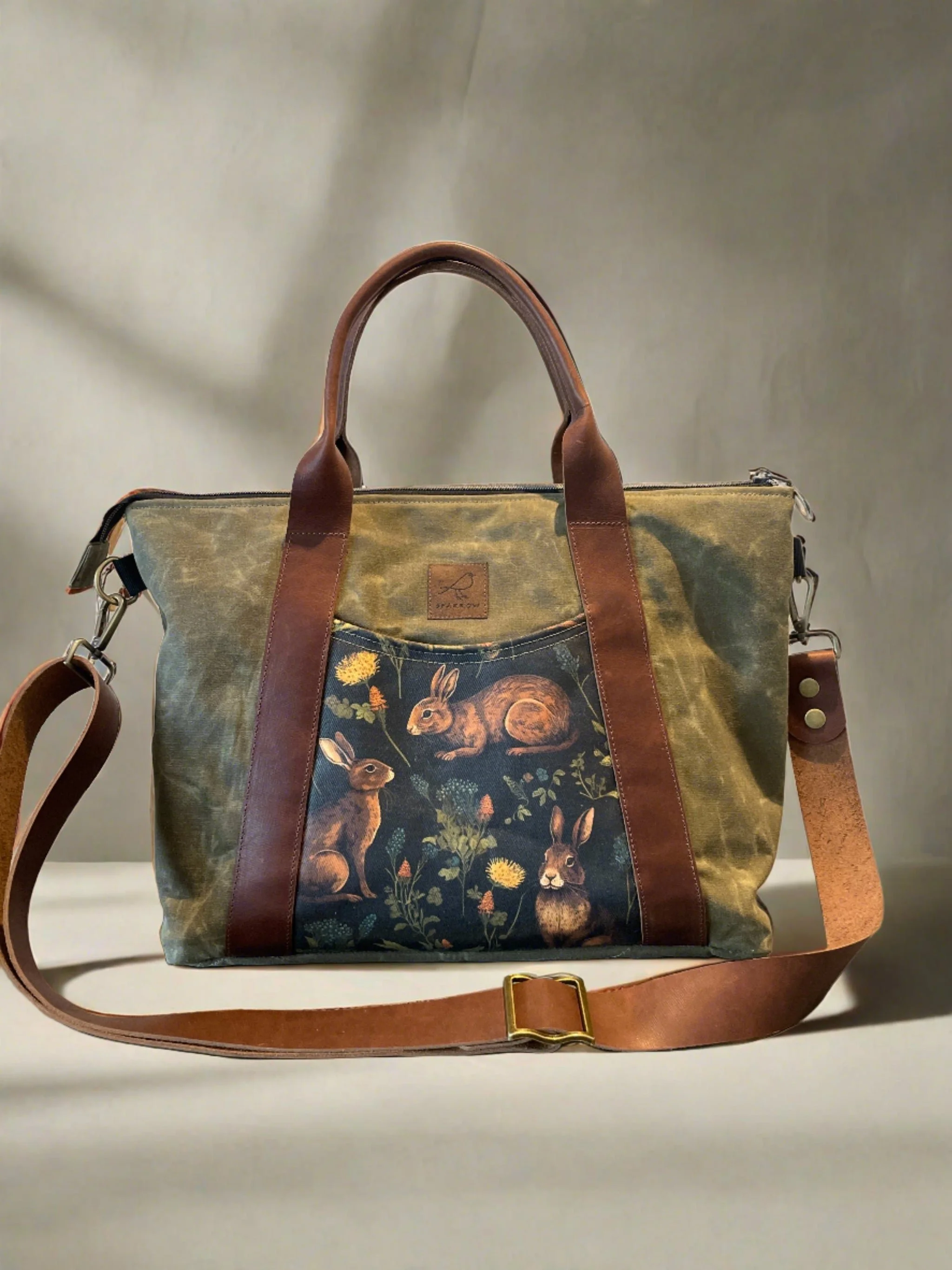 A handbag with a woodland animal print featuring rabbits and flowers, leather straps, and a detachable shoulder strap.
