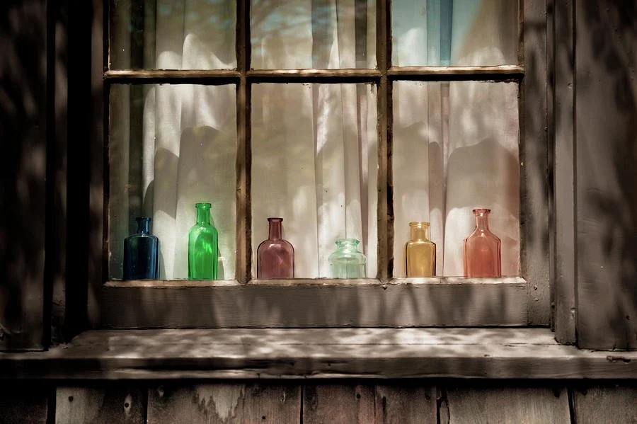 Six colorful glass bottles arranged on a windowsill inside an old wooden window, with sunlight casting shadows.