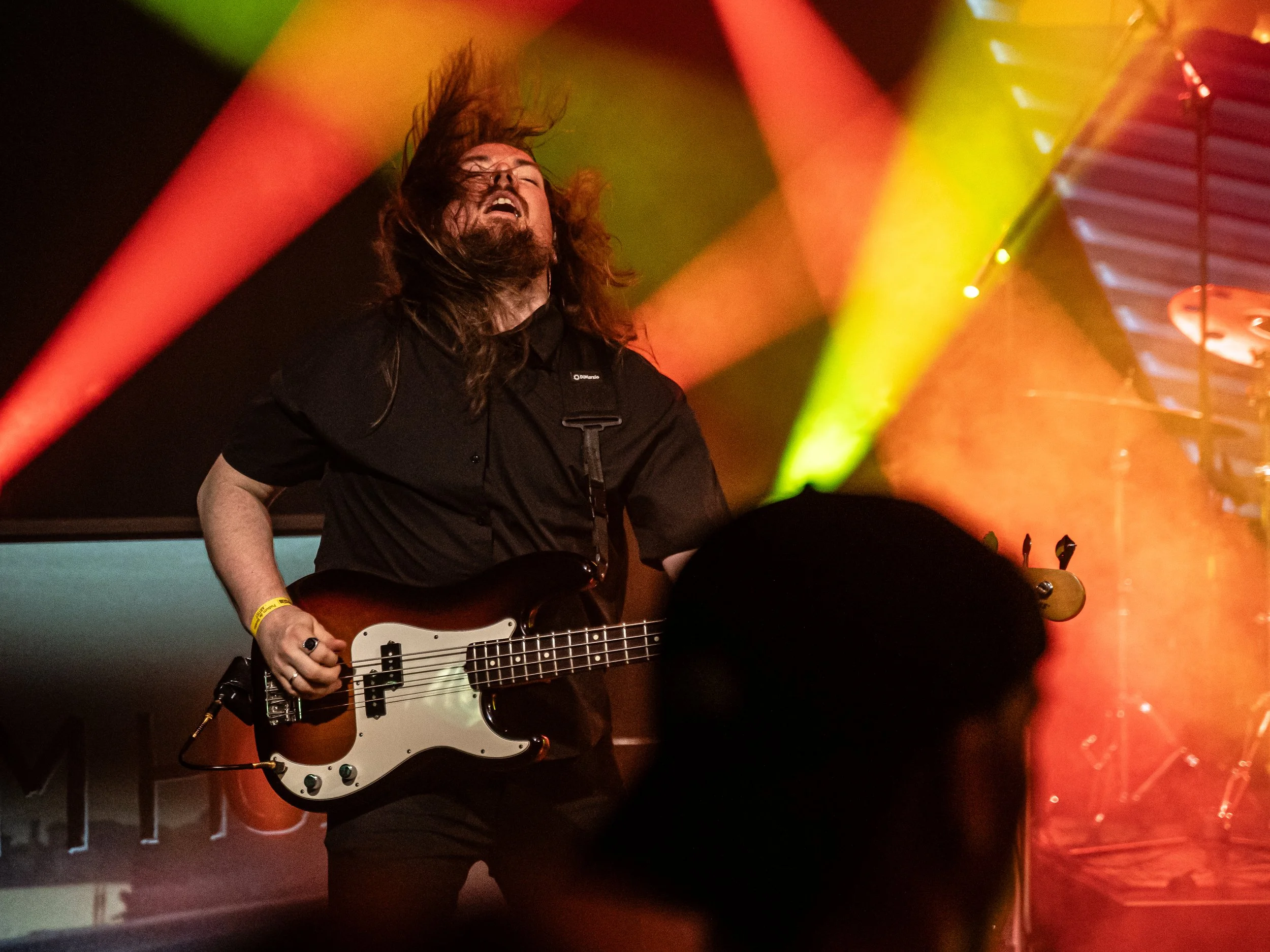 A long-haired musician playing bass guitar on stage with colorful stage lights and a drum set in the background.
