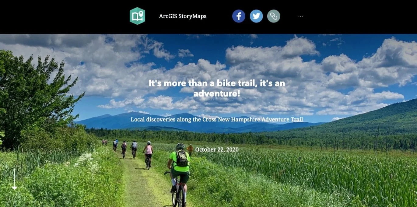 Photograph of a group of cyclists riding along a trail surrounded by green fields with mountains in the background under a partly cloudy sky.