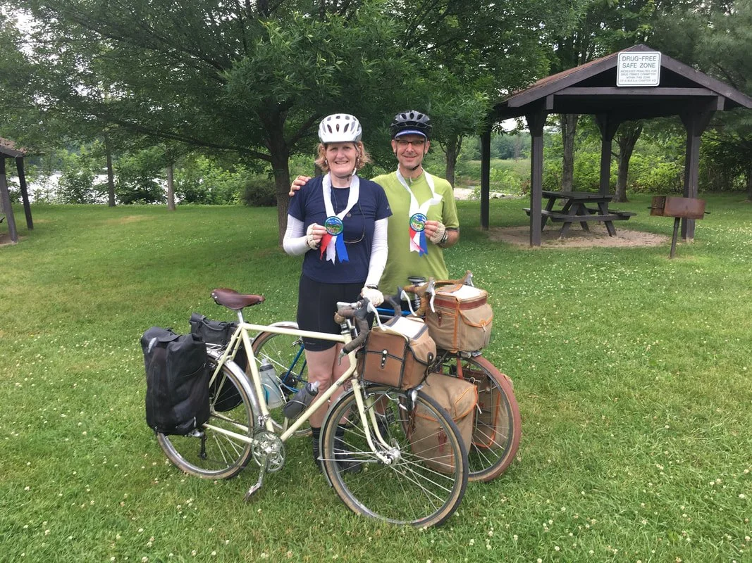 A smiling woman and man stand outdoors with their bicycles, wearing helmets and medals. The woman is holding a medal and standing next to a cream-colored bicycle with saddlebags, while the man is also holding a medal and standing next to a bicycle wi