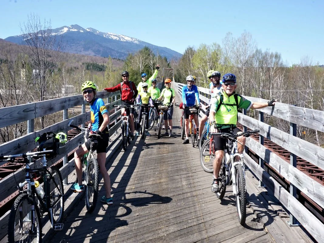 Group of people on bicycles posing on a wooden bridge with mountains in the background.