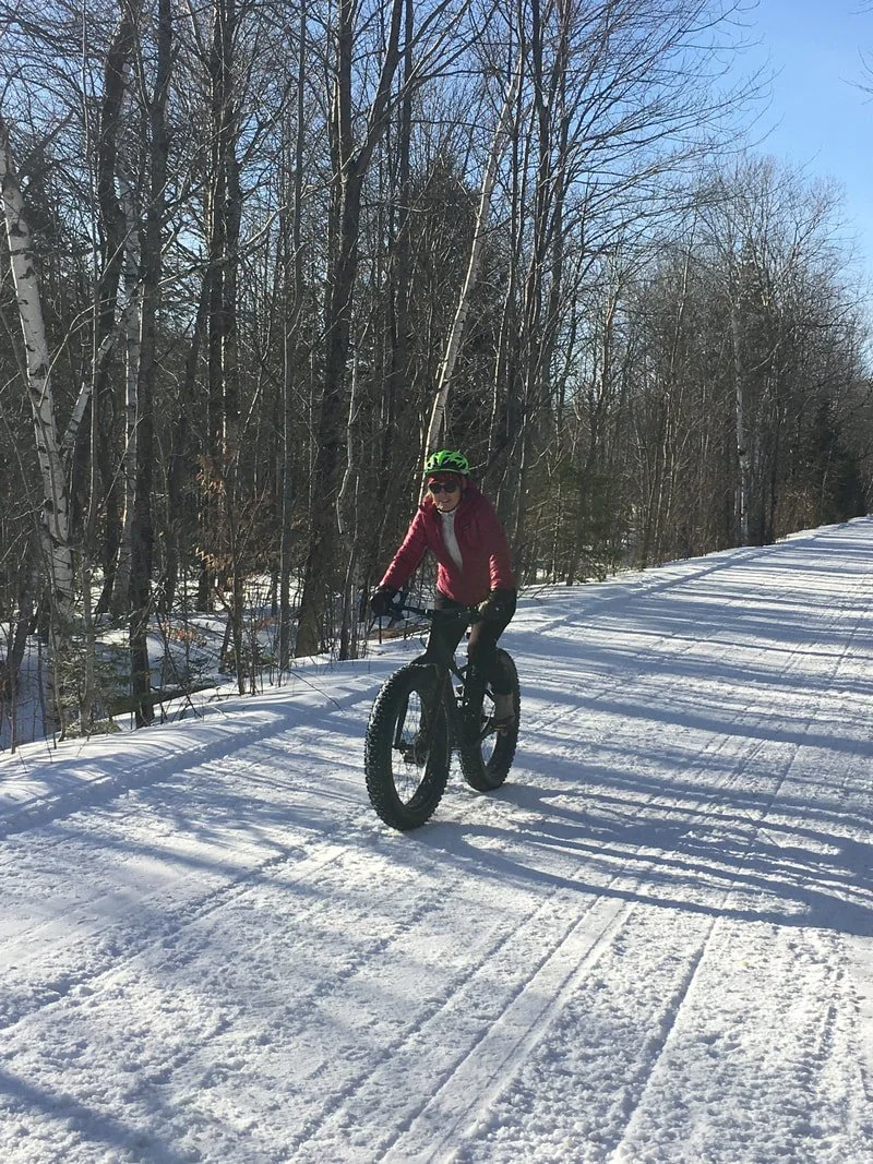 Person riding a fat tire bike on a snowy trail in a winter forest. The person is wearing a pink jacket, black helmet, and sunglasses, with leafless trees and clear blue sky in the background.