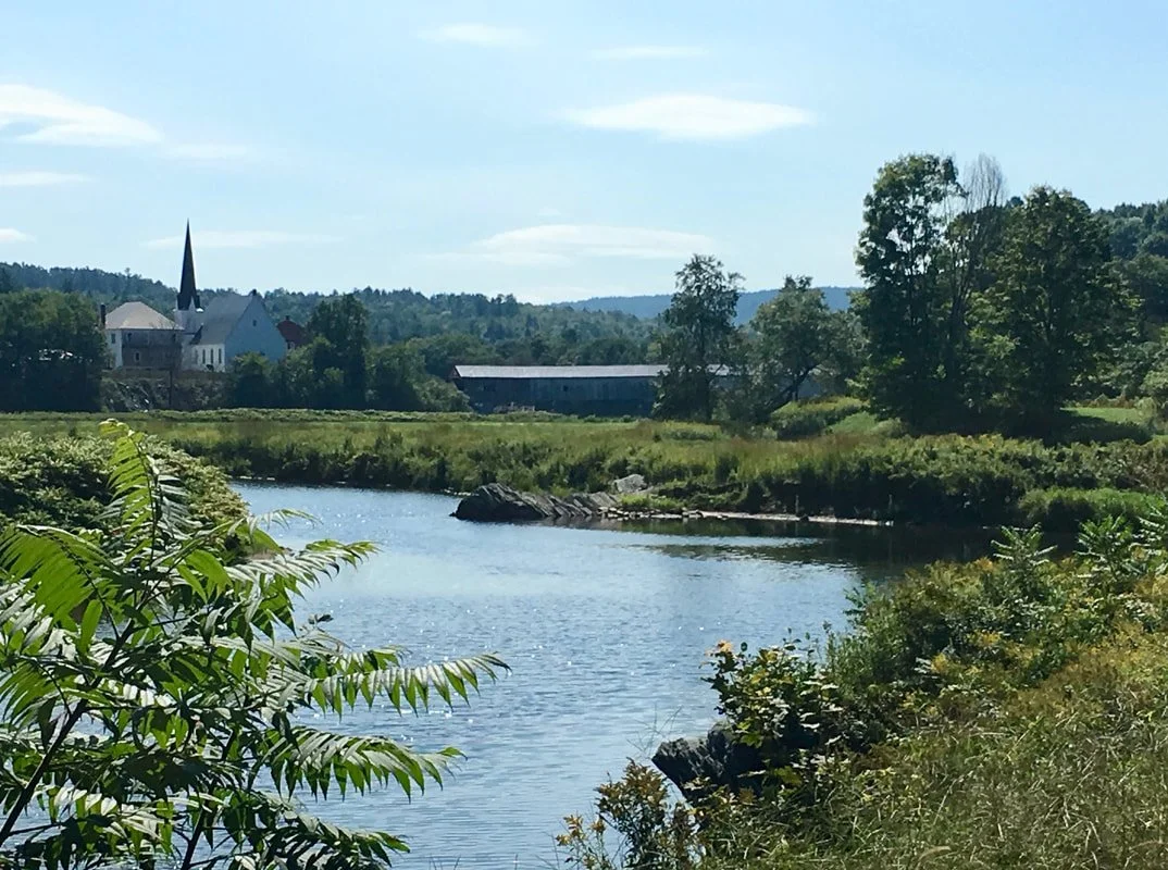A peaceful river scene with lush green trees and bushes along the banks, a distant church with a tall steeple, and low hills in the background under a partly cloudy sky.