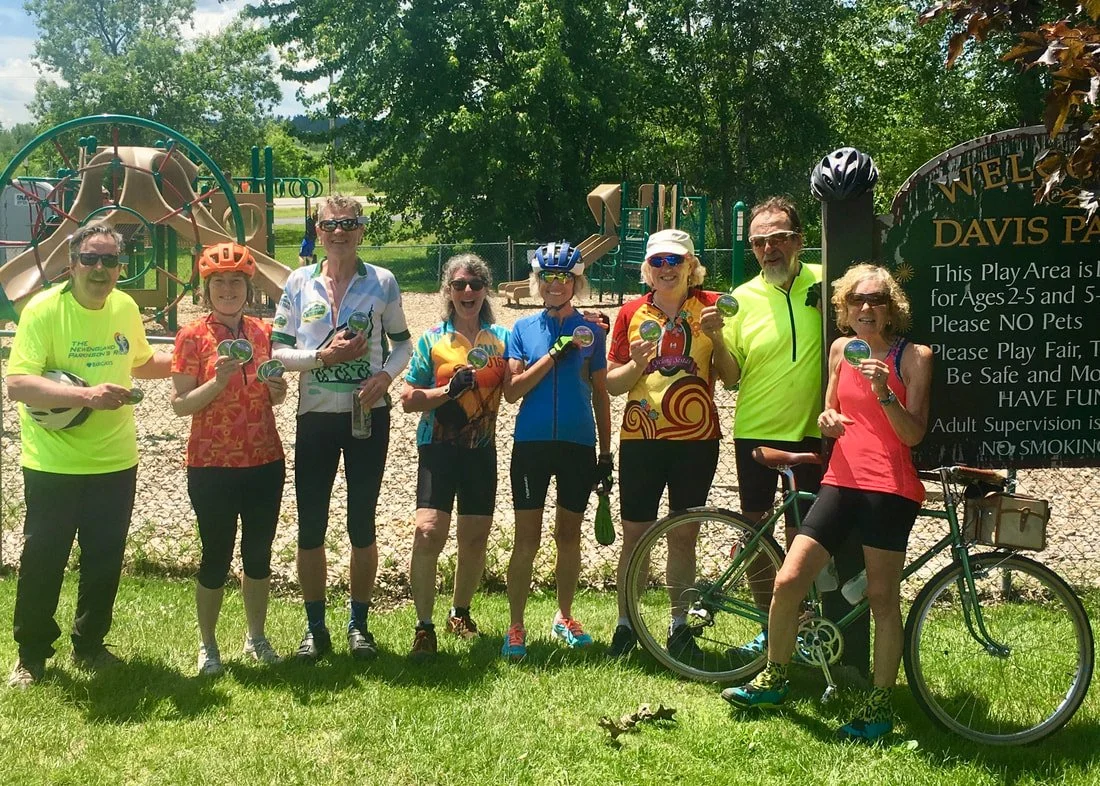 Group of nine people, mostly women, standing on grass in front of a playground at Davis Park, holding medals or awards. They are dressed in colorful activewear and cycling gear. One woman is standing next to a bicycle, and a sign indicates the playgr