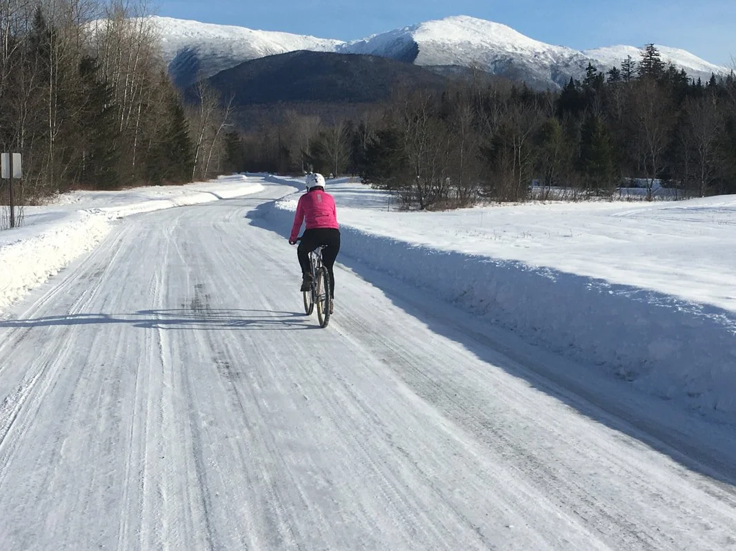 Person cycling on snow-covered road in winter landscape with mountains in the background.