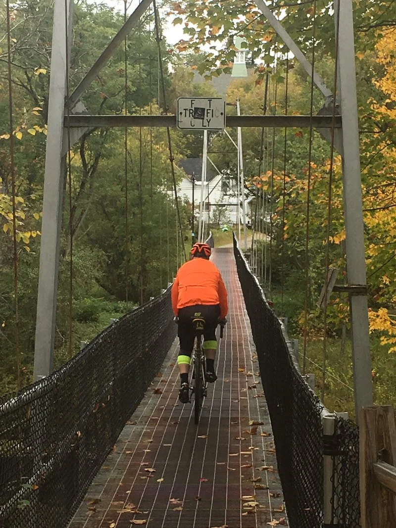 A person riding a bicycle across a narrow suspension bridge with safety netting on both sides, surrounded by trees with fall foliage.