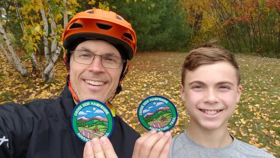 A man wearing glasses, an orange helmet, and a black jacket is holding a round patch that says 'Cross New Hampshire Adventure Trail' and a young boy with short brown hair in a gray shirt, both smiling outdoors during fall, with yellow leaves on the g