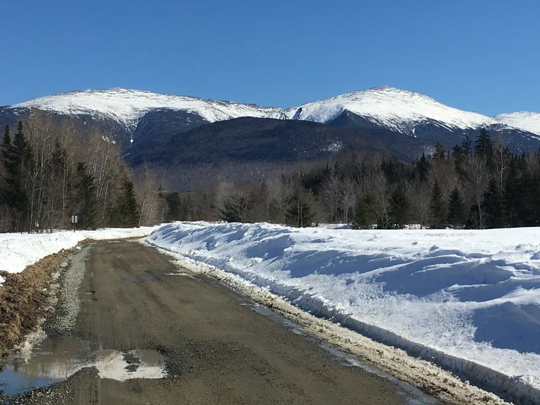 Snow-covered mountain landscape with a dirt road in the foreground.