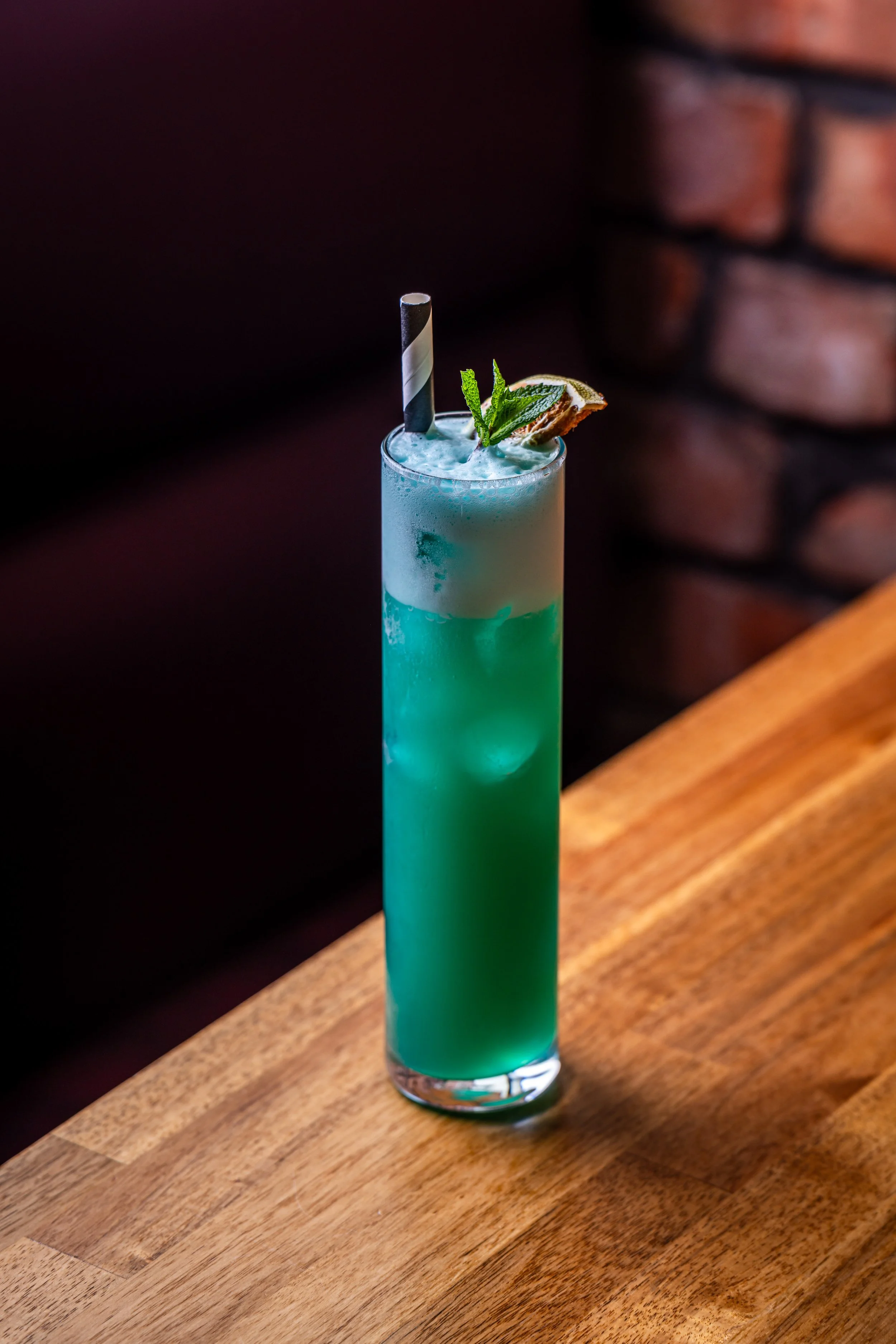 A tall glass containing a layered blue-green cocktail with ice, garnished with a lemon wedge, mint sprig, and a black-and-white striped paper straw, on a wooden surface against a dark background.