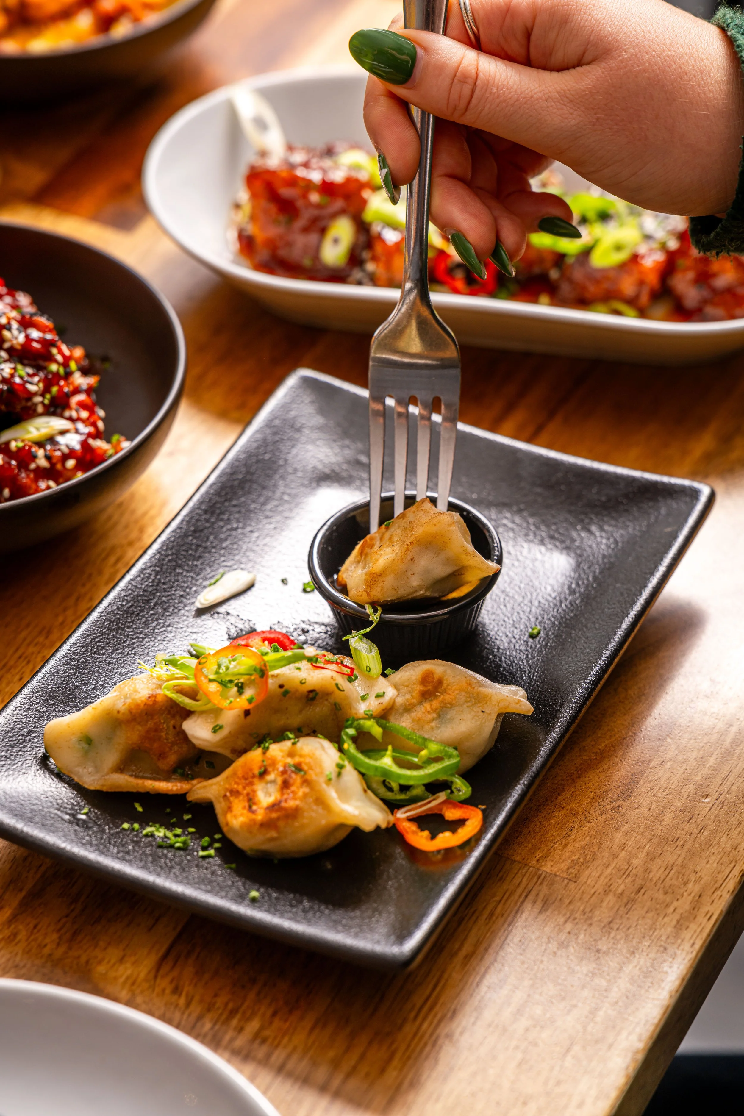 Close-up of a person with green nail polish holding a fork and picking up a dumpling from a black rectangular plate with three dumplings, garnished with chopped green onions and sliced chili, along with dipping sauce in a small black cup on a wooden 