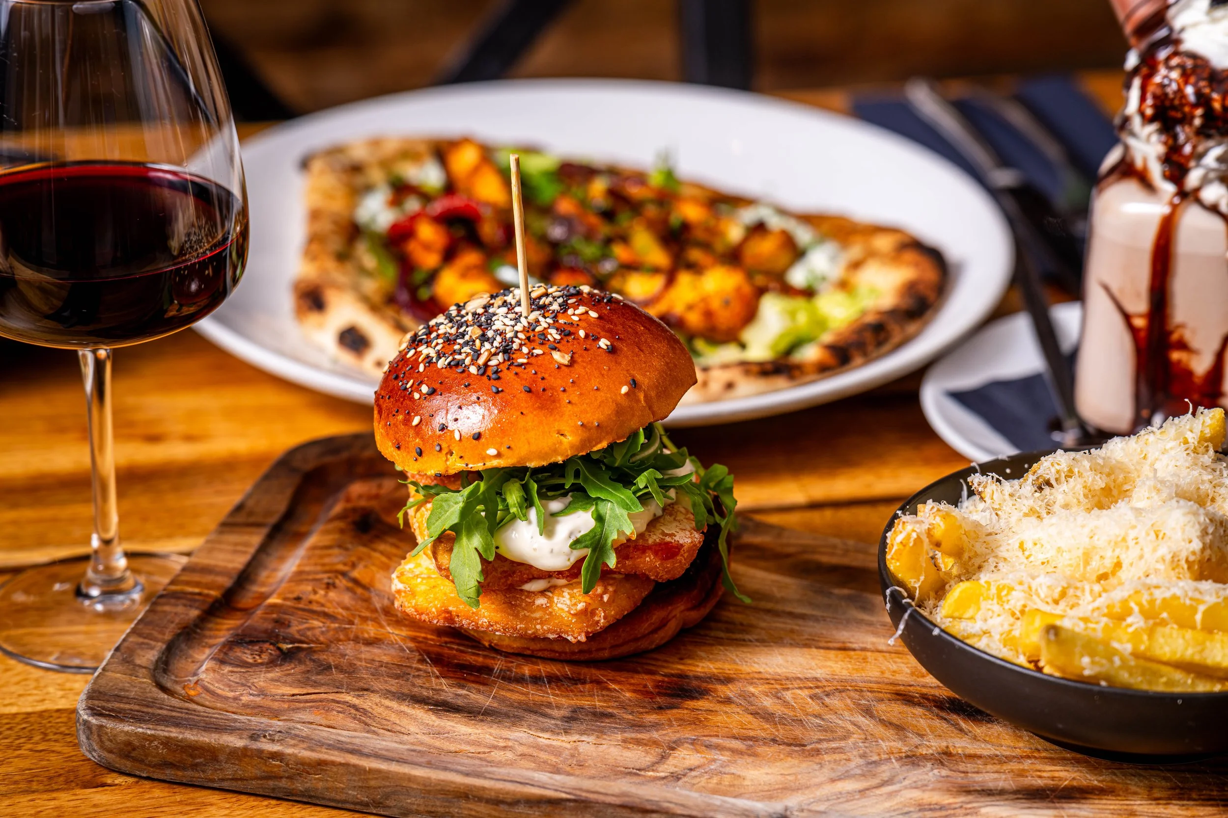 A meal featuring a burger with arugula, a fried fish fillet, and a brioche bun topped with sesame and black seeds, served on a wooden board. In the background, a glass of red wine, a plate of pizza, and a bowl of macaroni and cheese are visible.