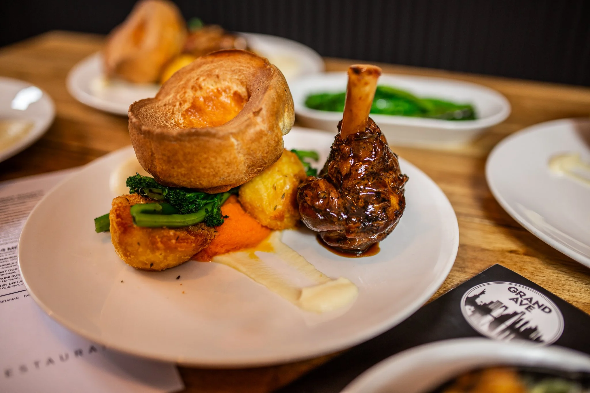 A plate with a Yorkshire pudding, green vegetables, a piece of roasted chicken with gravy, and some roasted potatoes, on a wooden table.