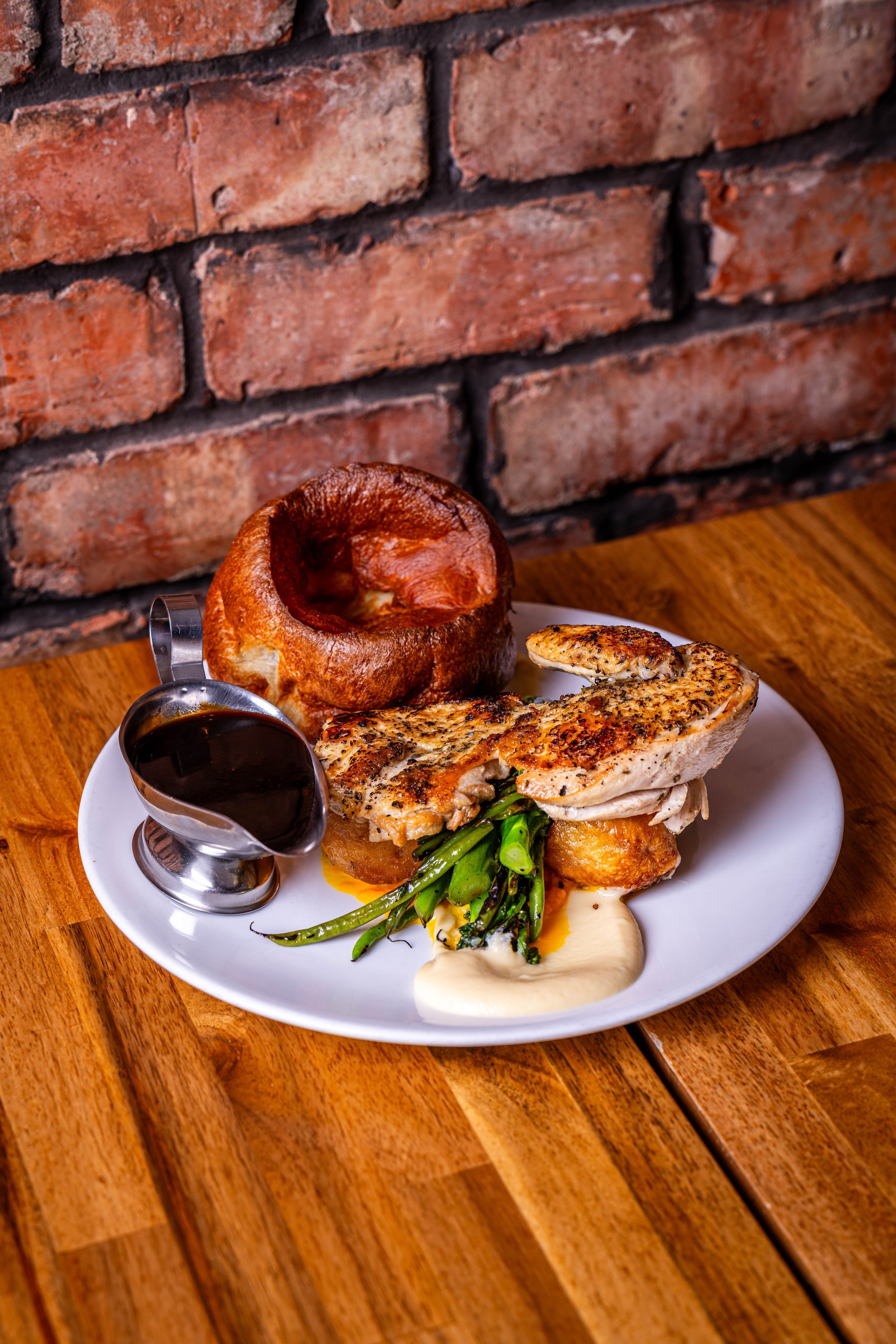 Plate with fried chicken, green beans, mashed potatoes with gravy, bread roll, and a small pitcher of dark gravy on a wooden table with a brick wall background.