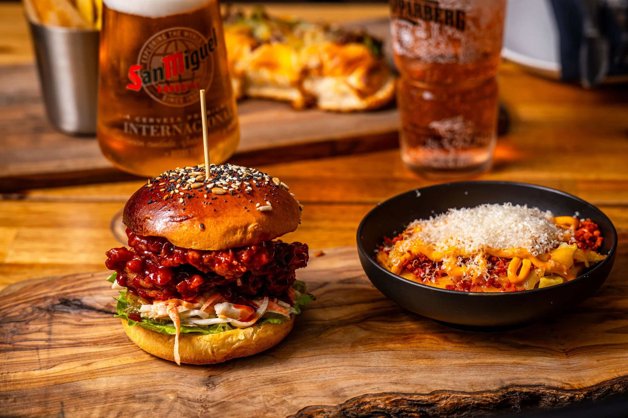 A burger with a sesame seed bun, fried chicken fillet, lettuce, coleslaw, and sauce on a wooden serving board, with a bowl of pasta with ground meat, cheese, and vegetables beside it. In the background, there are two glasses of beer, one clear and on