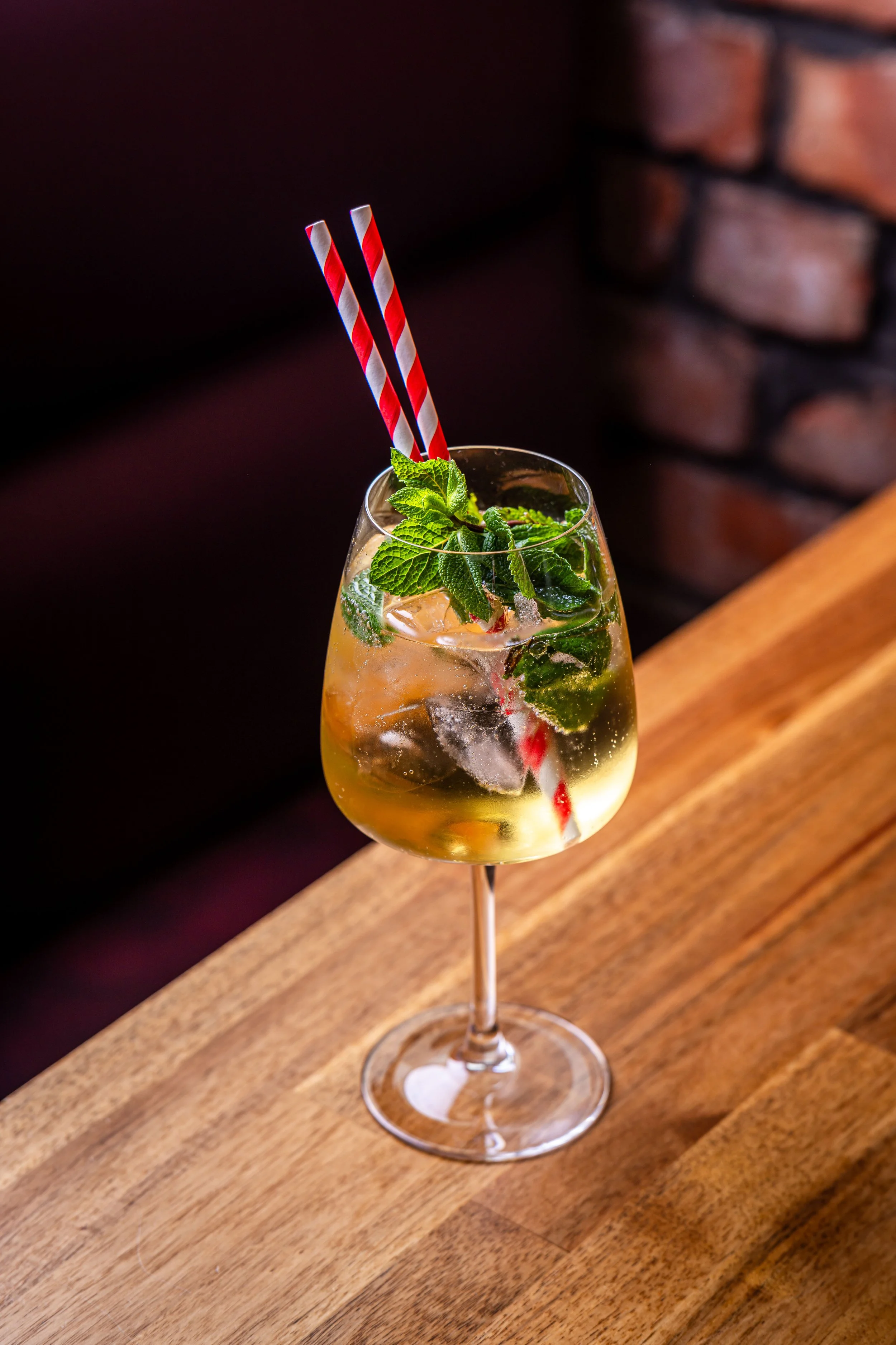 A cocktail in a wine glass with ice, fresh mint leaves, and two red and white striped paper straws on a wooden surface.
