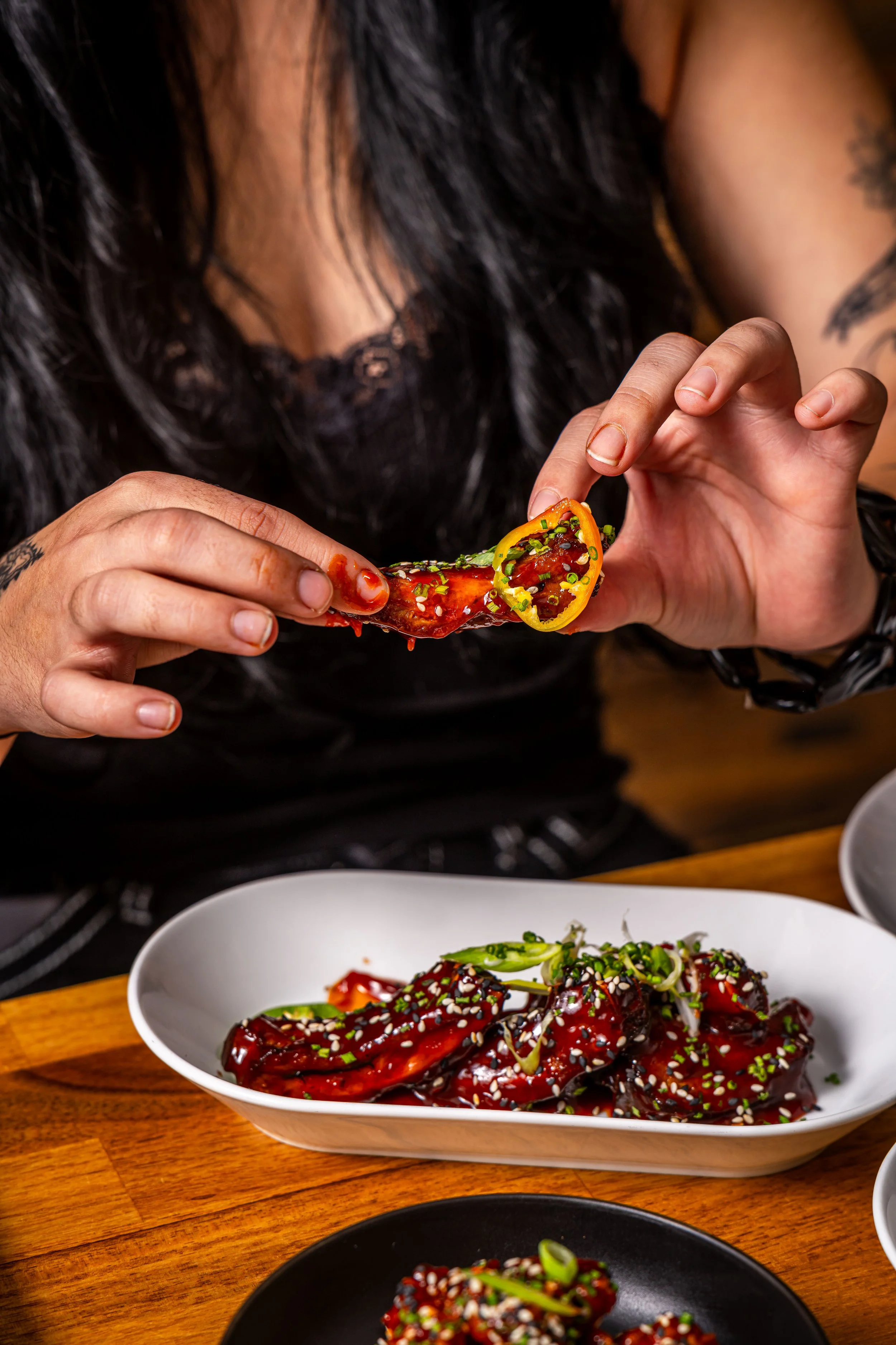 Person holding a piece of glazed Asian-style chicken wing garnished with chopped green onions over a plate of similar chicken wings, some garnished with sesame seeds, on a wooden table.