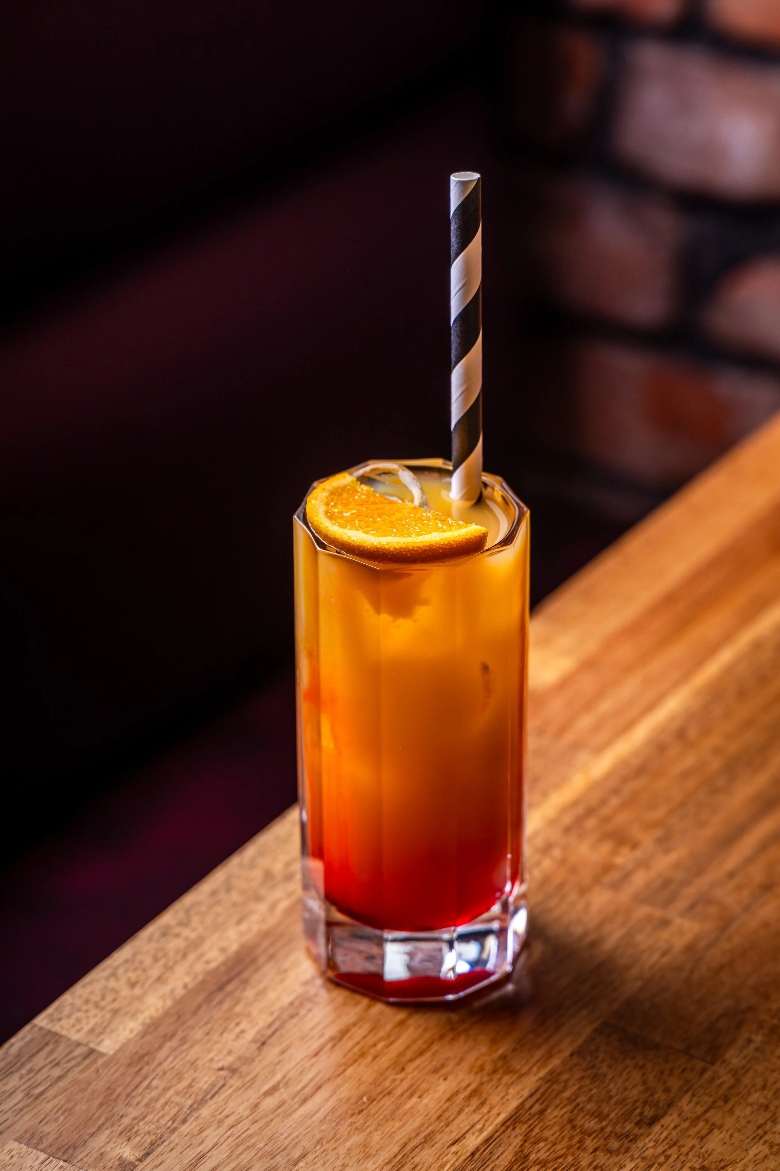 A tall glass of layered cocktail with orange and red hues, garnished with an orange slice and a striped straw on a wooden bar counter.