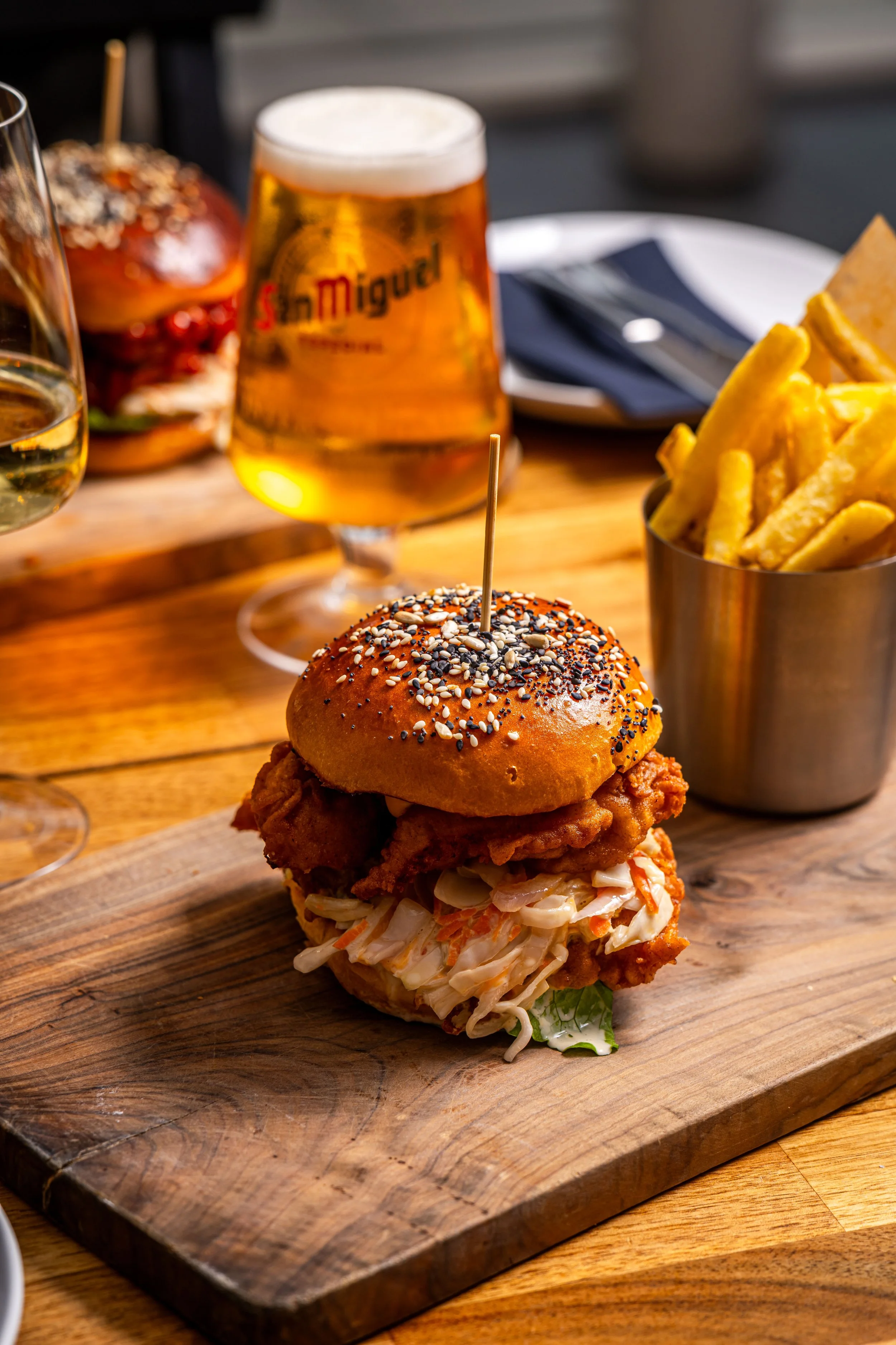 Fried chicken sandwich with coleslaw on a bun, served on a wooden board with a side of French fries and a glass of beer.