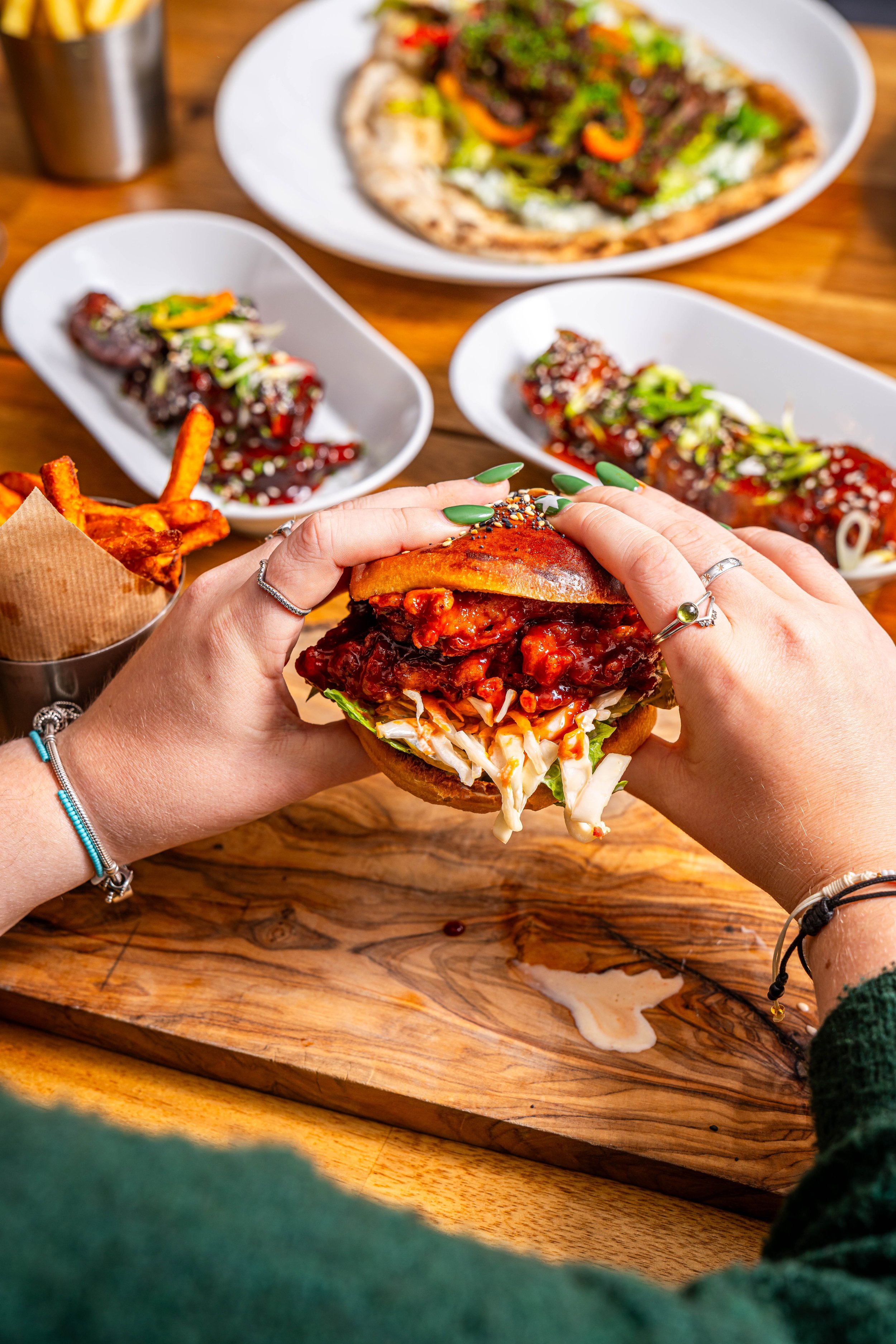 Person holding a barbecue pulled pork sandwich with coleslaw, surrounded by bowls of other Asian-inspired dishes and fries on a wooden table.