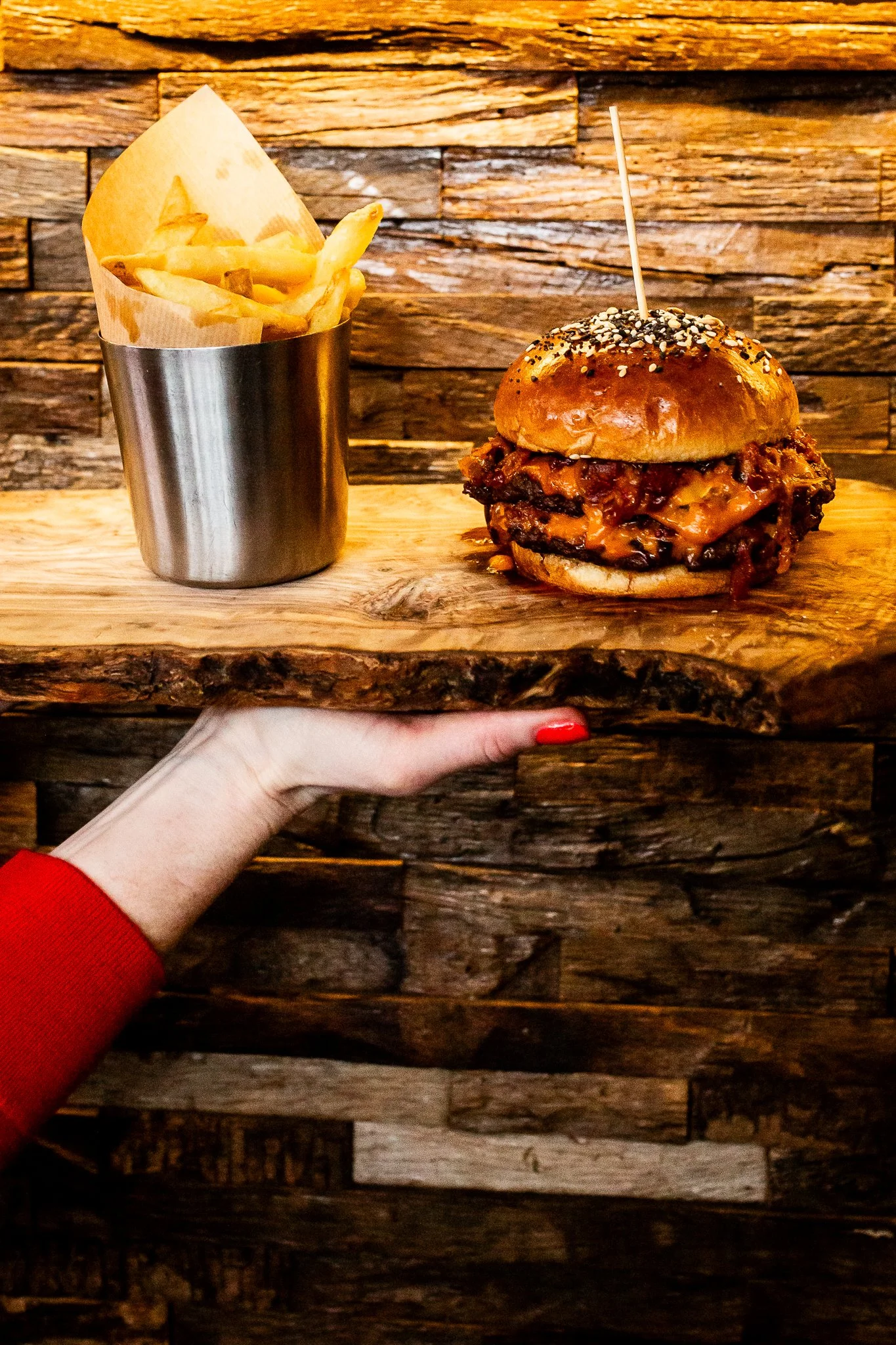 A hand with red painted nails holds a wooden board with a burger topped with sesame seeds and a skewer, alongside a metal cup of French fries lined with parchment paper, against a rustic wooden wall background.