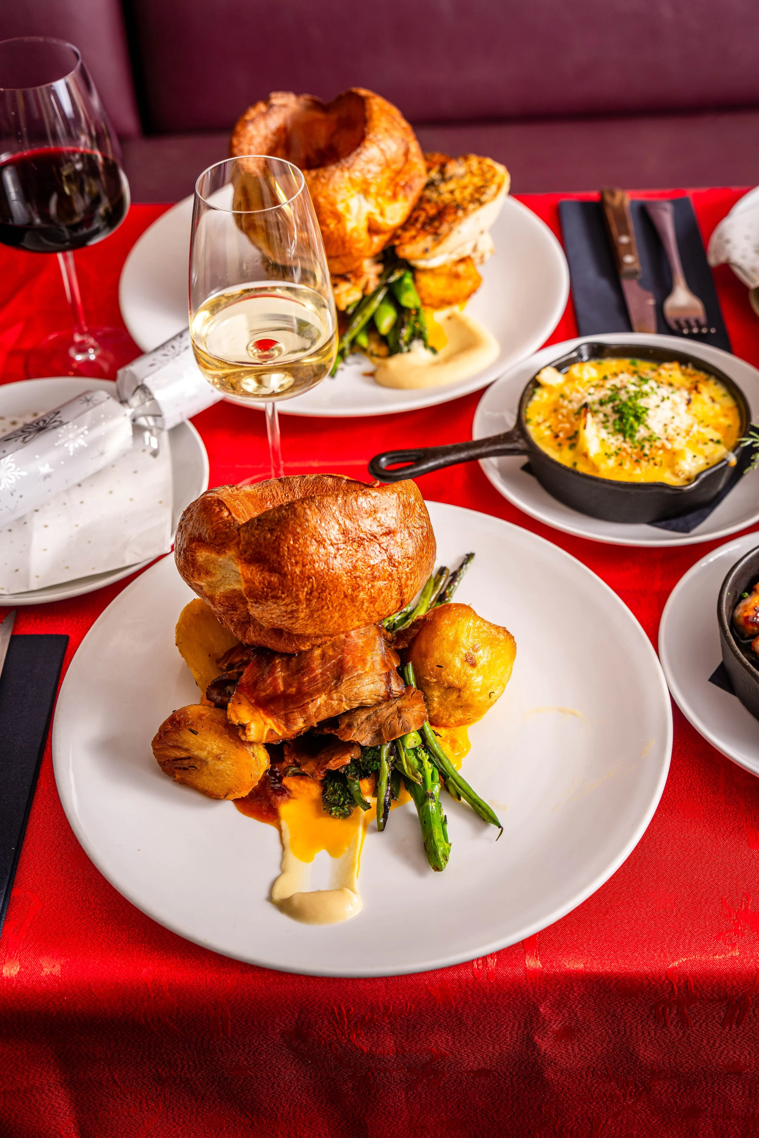 A dining table with a red tablecloth, set with two glasses of white and red wine, a wrapped gift, and multiple dishes including a feast with roast chicken, vegetables, and side dishes.