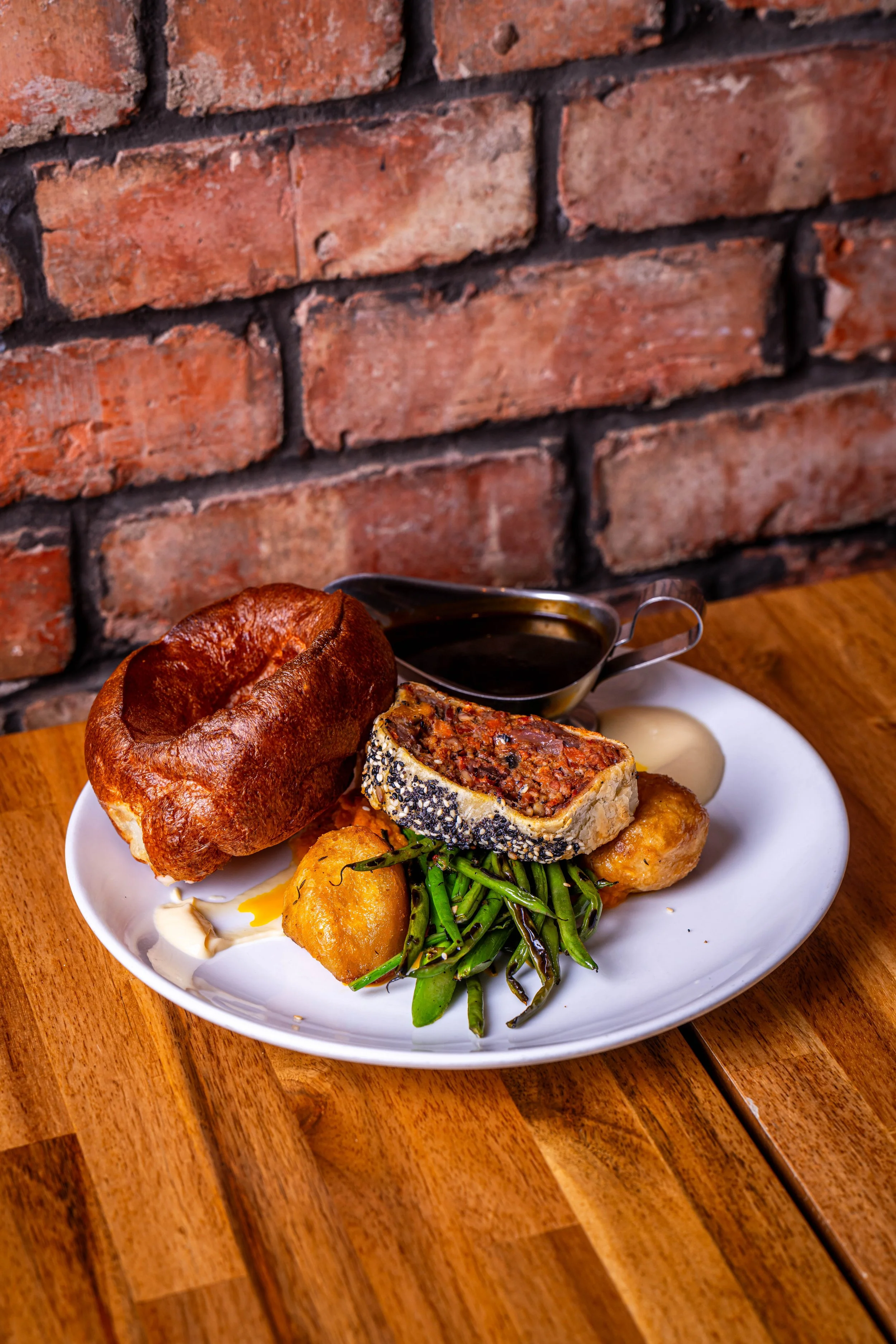 Plate with bread, beef Wellington, green beans, hush puppies, sauce, and mayonnaise, on a wooden table against a brick wall.