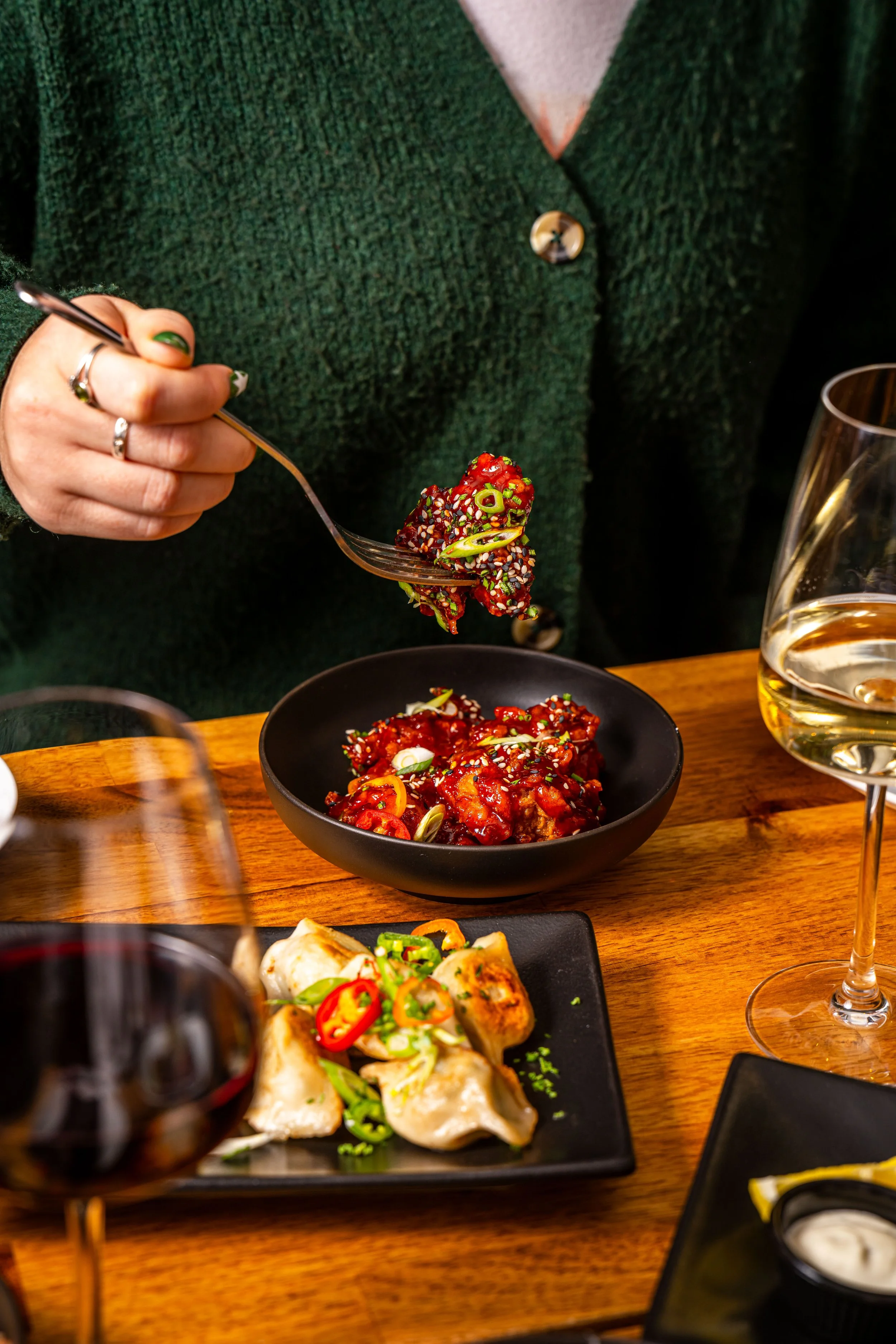 A person in a green sweater is holding a fork with a serving of spicy Korean barbecue beef, surrounded by dishes of food and glasses of white wine on a wooden table.